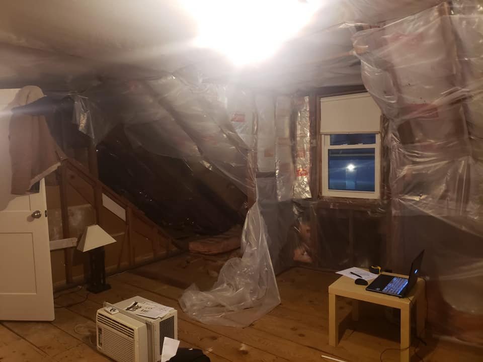 Room under renovation with plastic sheeting covering walls, a window, and ceiling; small table with laptop, desk lamp, and air conditioning unit on floor.