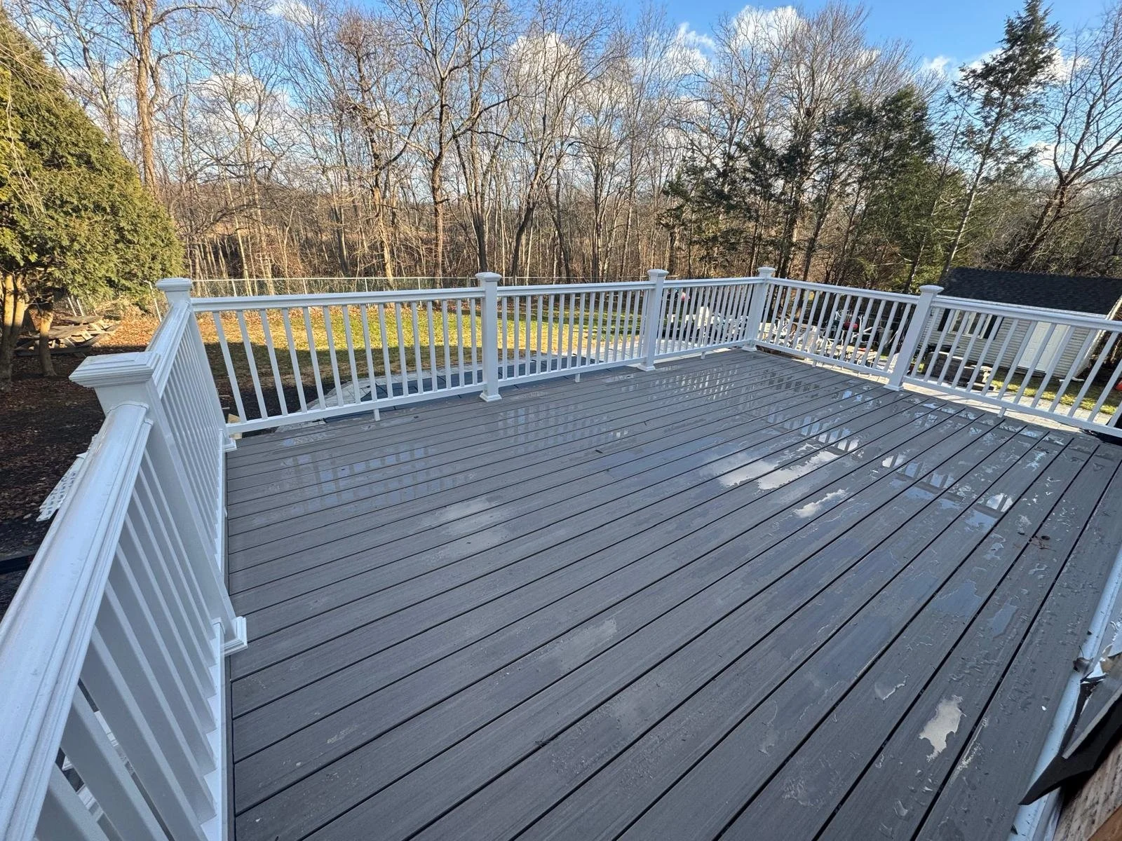 Empty outdoor deck with wet dark gray wooden planks and white railing, overlooking a backyard with trees and a neighboring house.