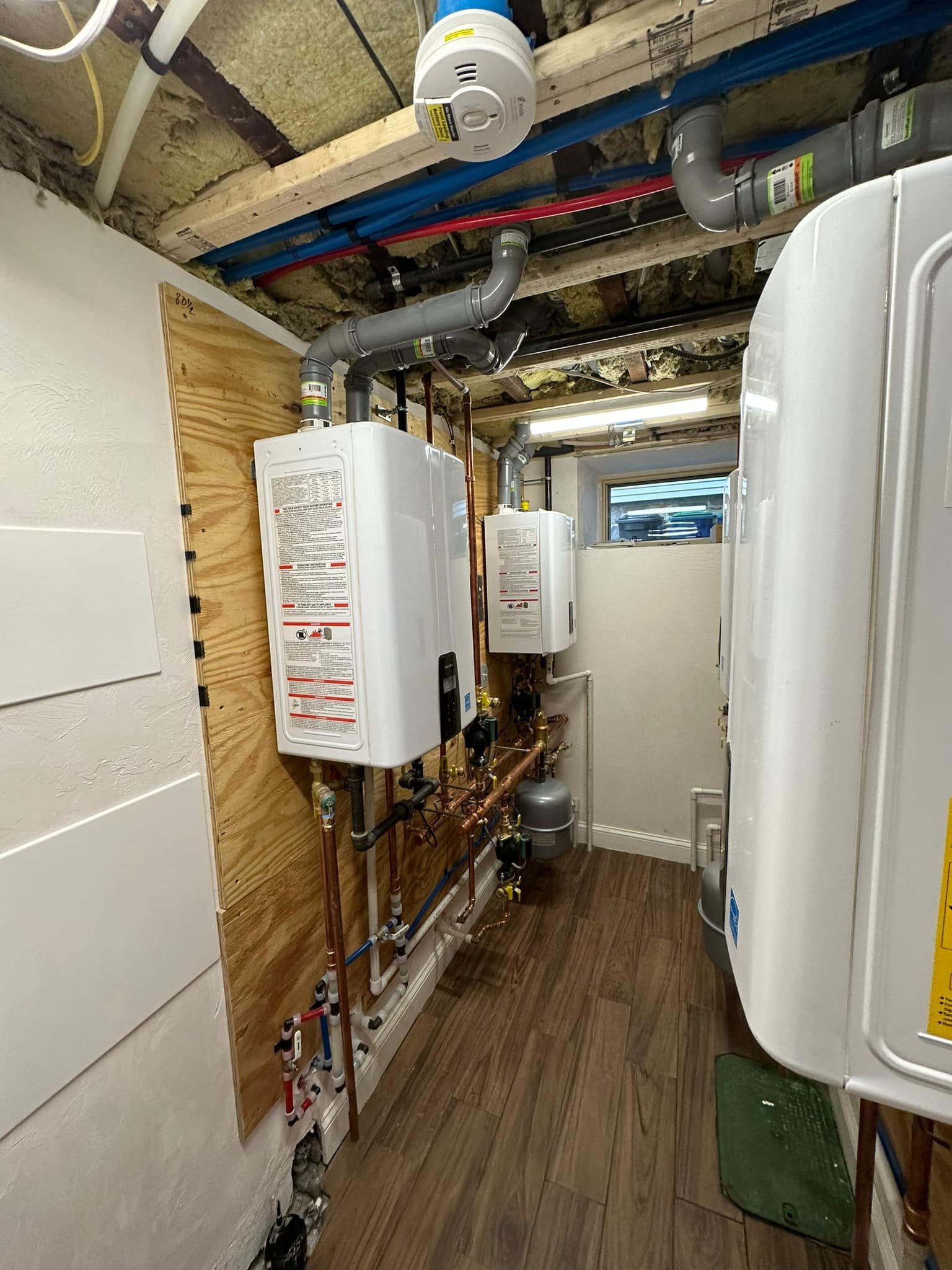A utility room with two white tankless water heaters mounted on a wooden wall, with pipes and valves below them. The ceiling is unfinished with insulation and pipes, and there is a small window in the back of the room.