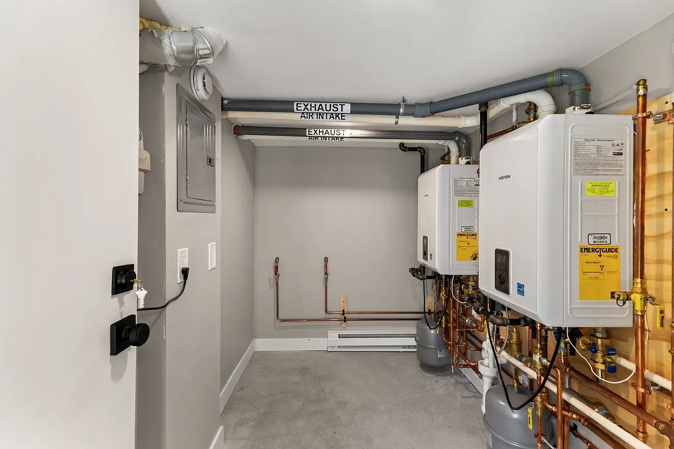 A utility room with copper pipes, two white tankless water heaters, gray drain pipes, and labels for exhaust and air intake.