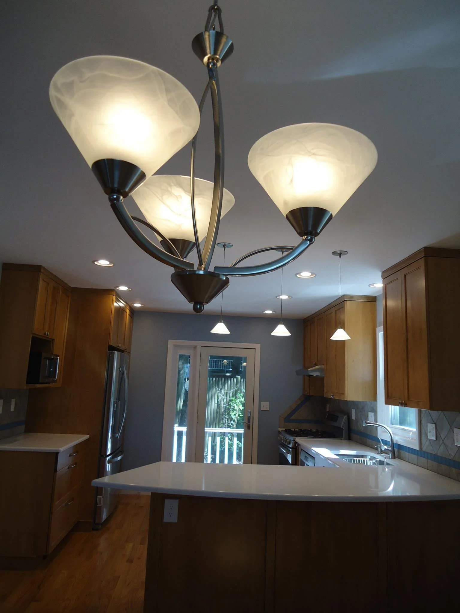 A modern kitchen with a large ceiling light fixture featuring three frosted glass lampshades and a stainless steel frame. The kitchen includes wooden cabinets, stainless steel appliances, a white countertop island, and a sliding door leading outside.