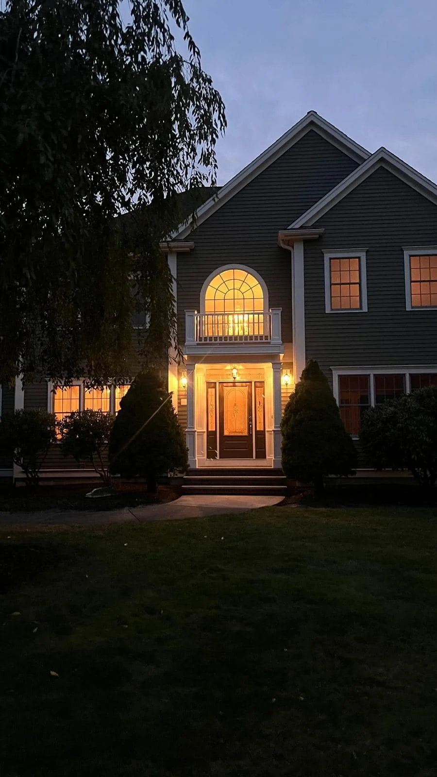 A two-story house at dusk with warm lighting inside, large arched window above the front door, surrounded by bushes and a well-kept lawn.