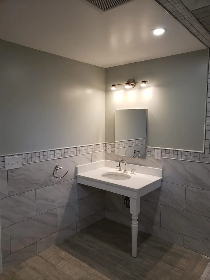 A bathroom with a white corner sink, gray walls, tile wainscoting, and a small mirror. Overhead lighting is mounted above the mirror, and there is a towel ring on the wall to the left of the sink.