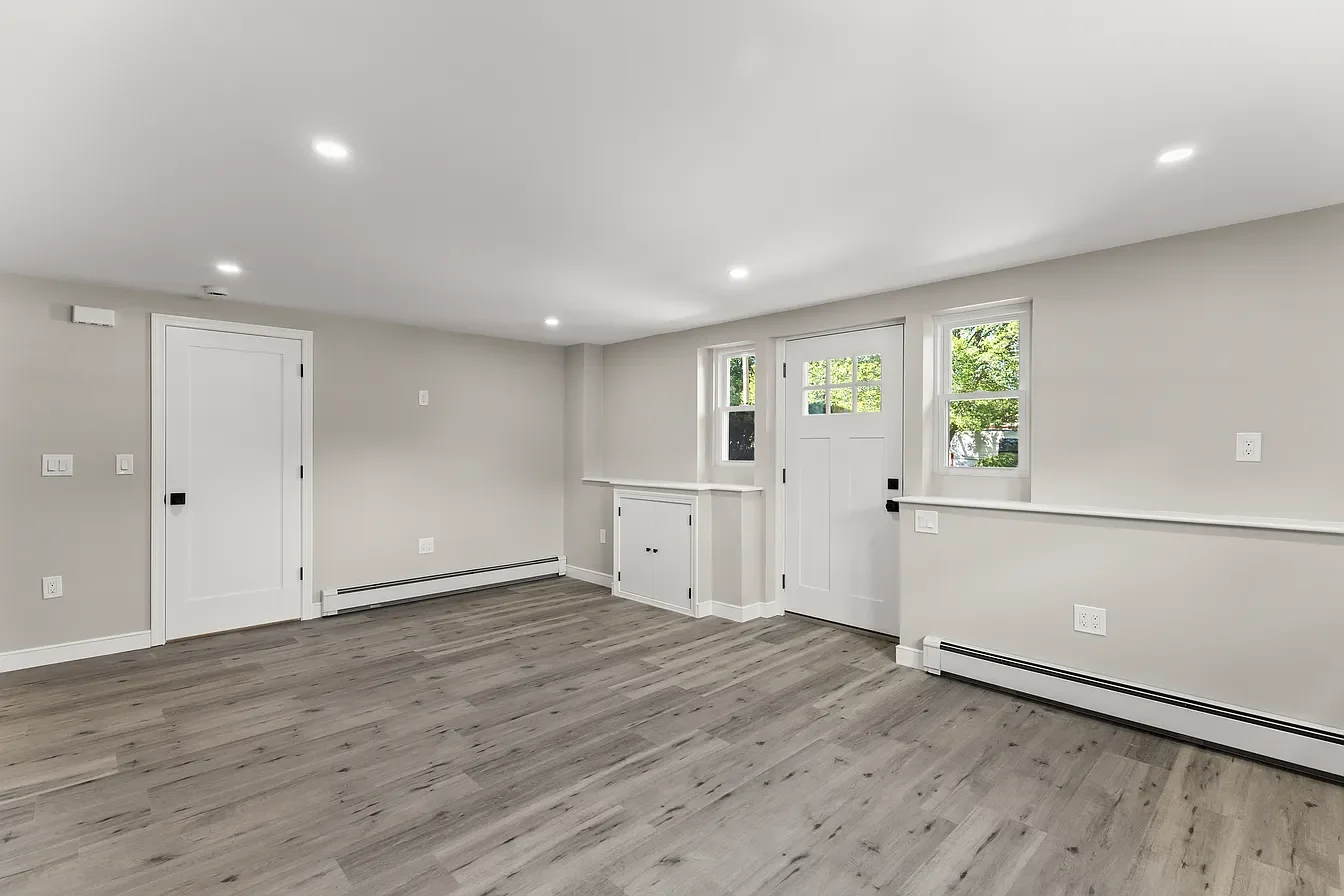An empty living room with light gray walls, white trim, three small windows, a white door with a window, and wood-look flooring.
