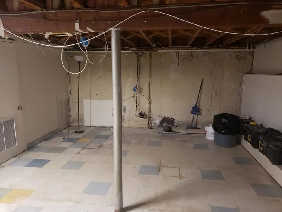 A basement with exposed ceiling beams, a metal support post, unfinished wall with plumbing, and tools including a vacuum and a toolbox.