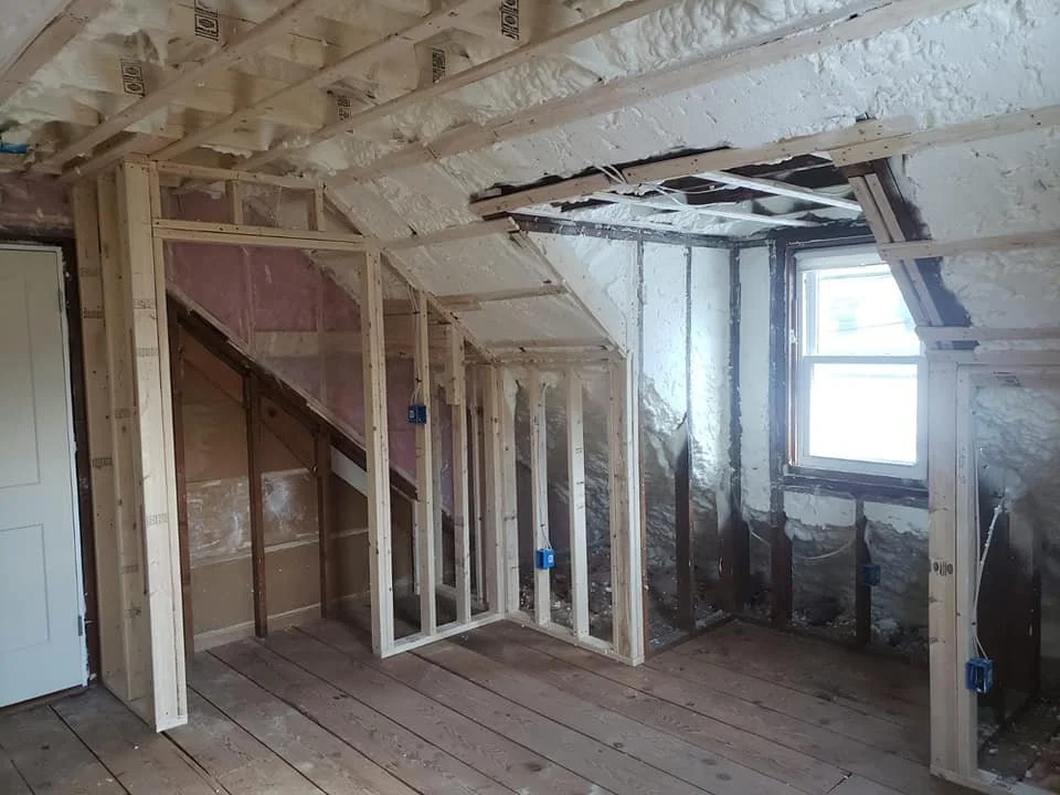 Interior of an attic or upper room under construction with exposed wooden framing, wiring boxes, a window, and partially finished walls.