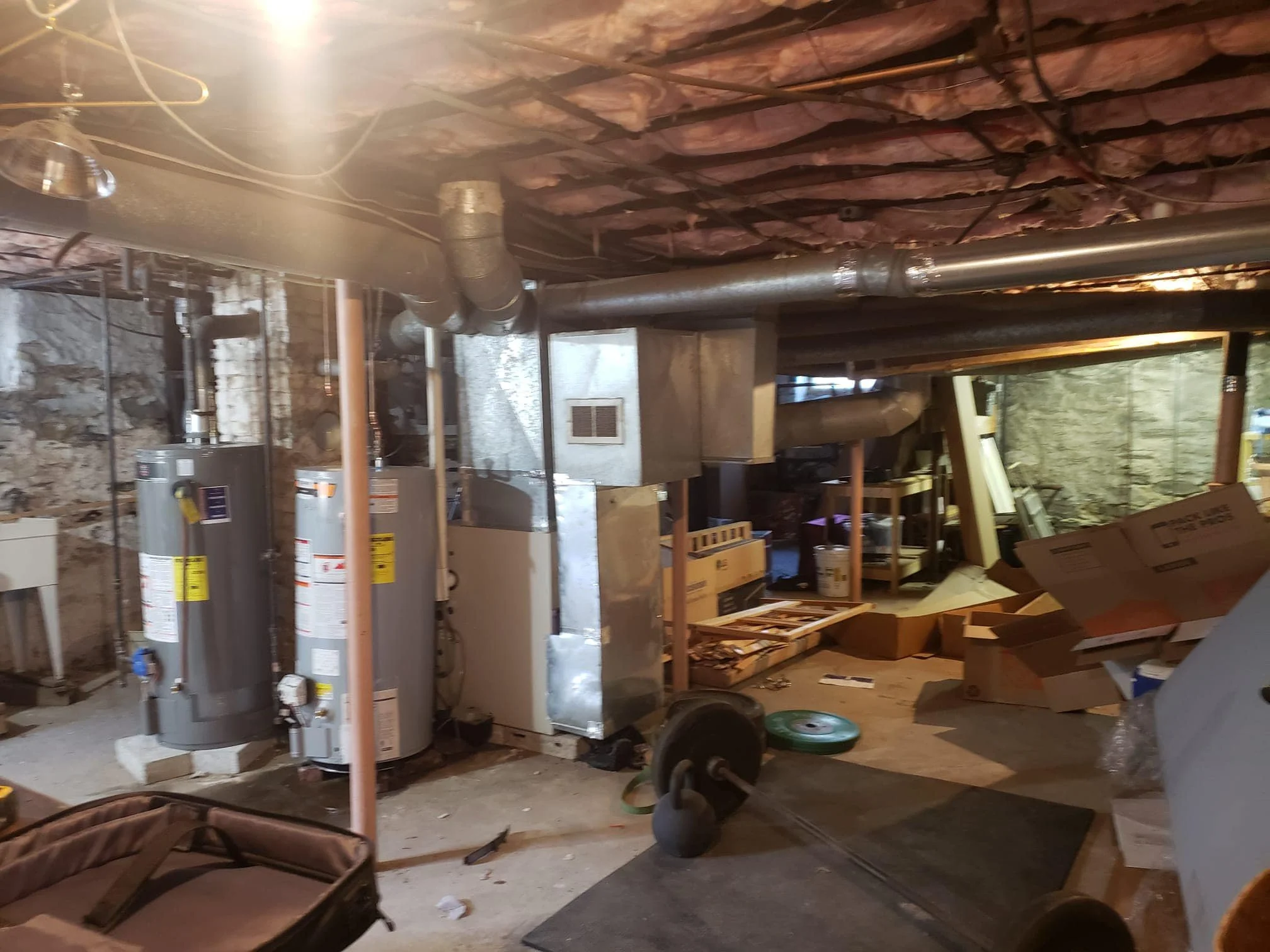 Inside a basement with exposed insulation and plumbing, showing water heaters, an HVAC system, weights, a kettlebell, cardboard boxes, and various stored items.