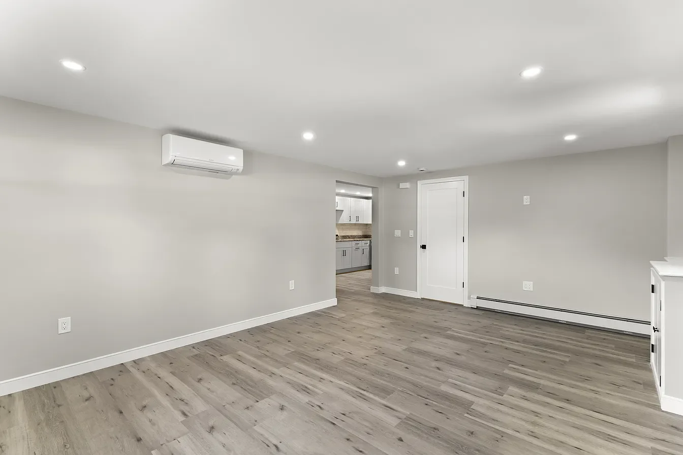 Empty living room with light gray walls, white trim, and wood-look flooring, with visible kitchen through doorway.