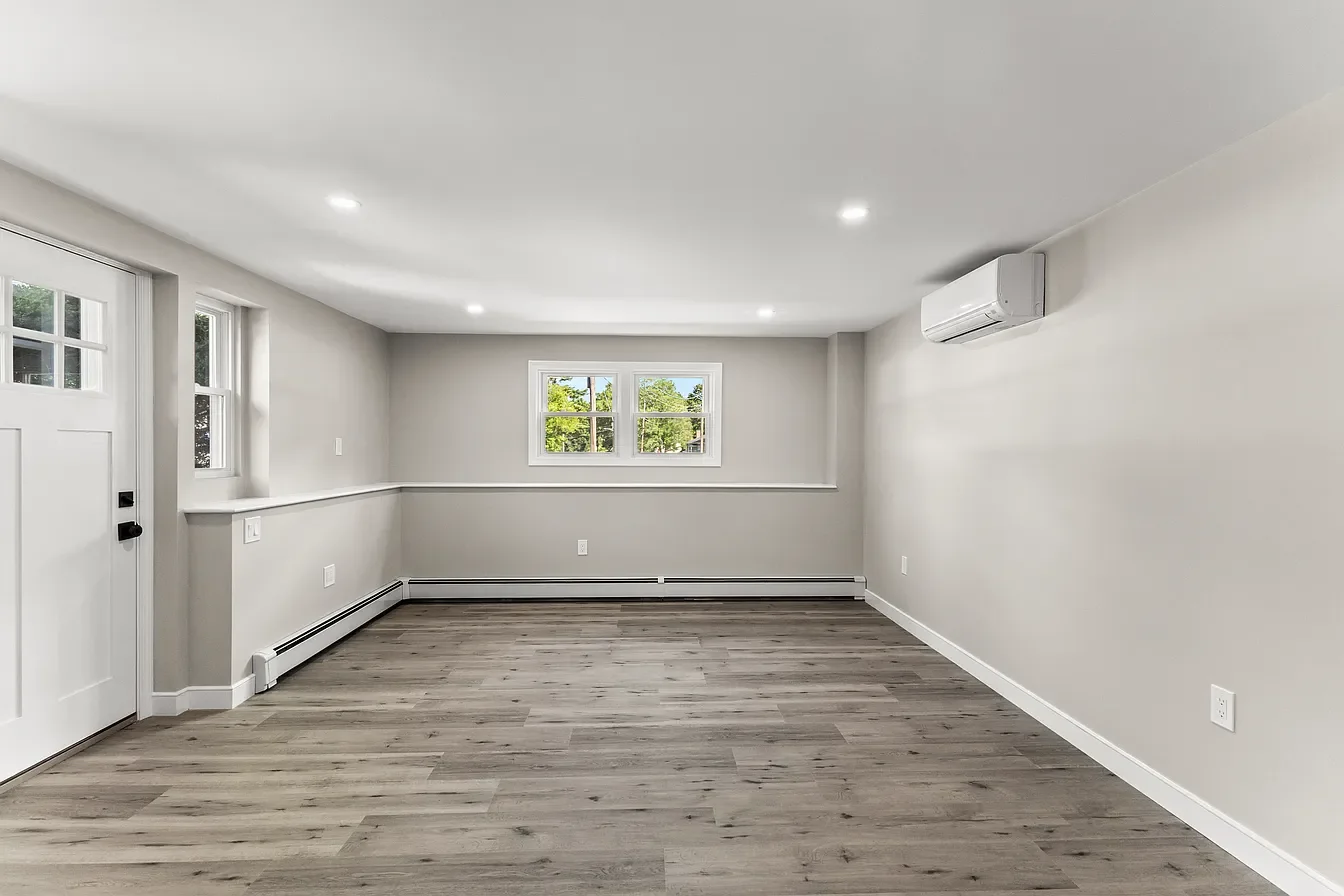 Empty living room with light gray walls, wood flooring, and three windows, with ceiling lights and a wall-mounted air conditioner.
