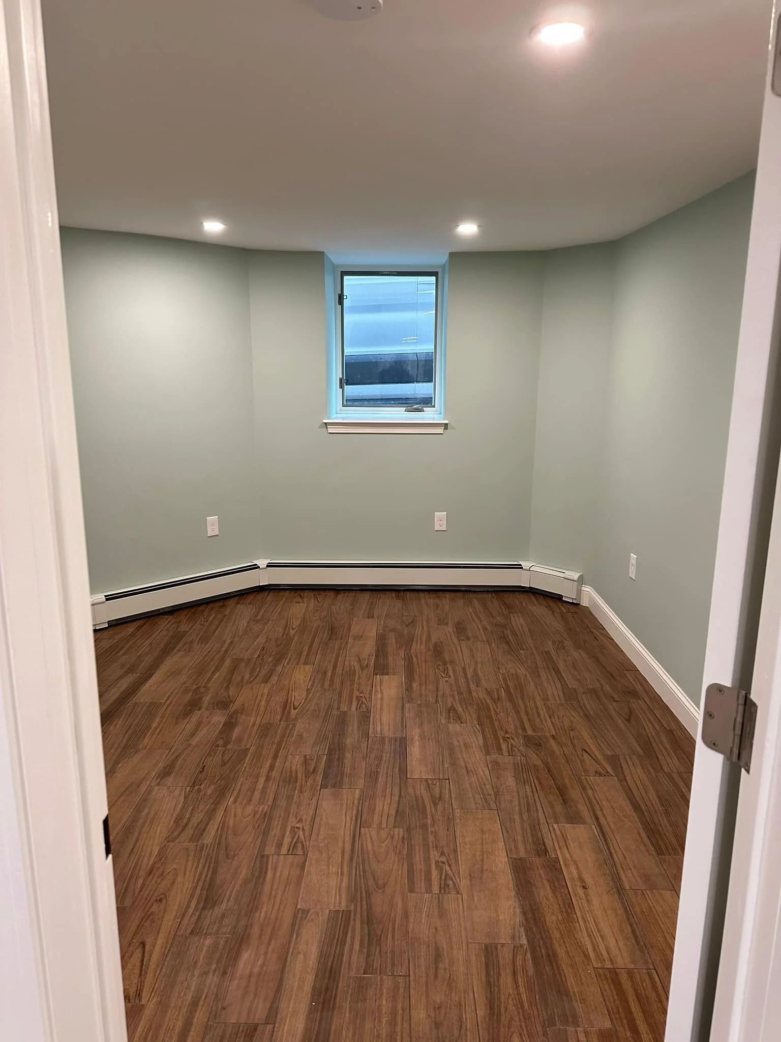 Empty room with light green walls, a small window, hardwood flooring, and ceiling lights.