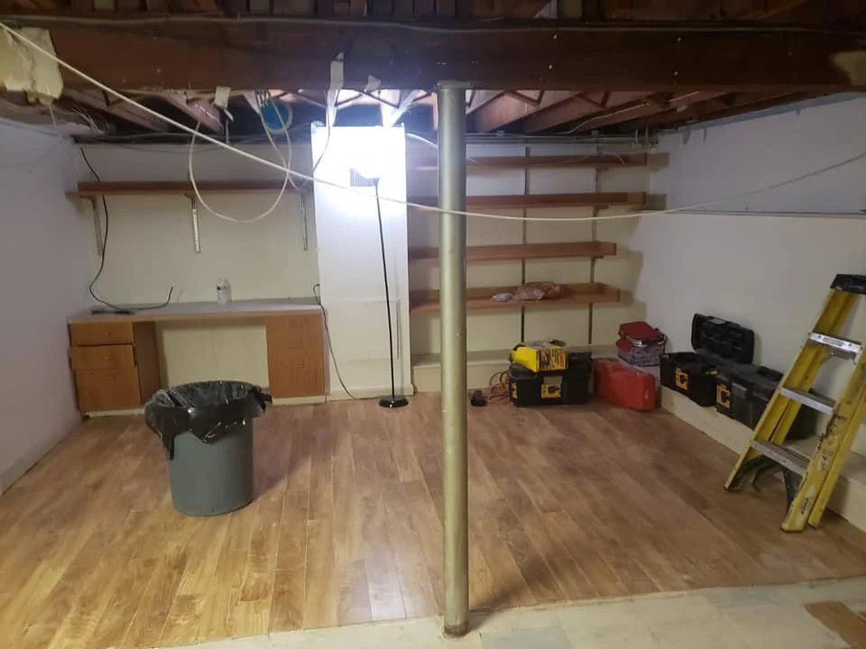 Empty room with wooden floor, unfinished ceiling, a trash can, a ladder, and several power tool cases and shelves along the walls.