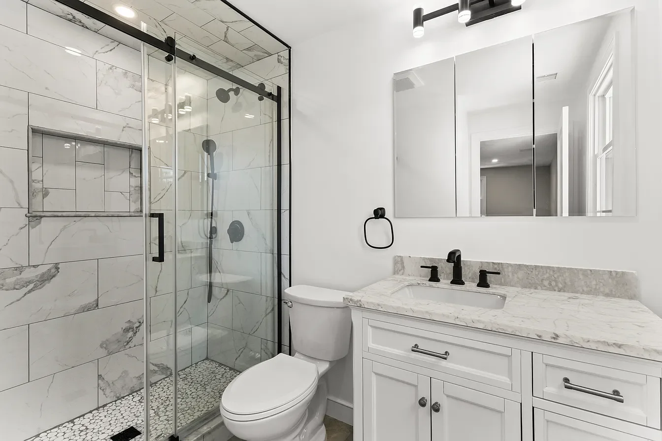 Modern bathroom featuring a glass-enclosed marble shower, a white toilet, a white vanity with a marble countertop, a wall-mounted mirror, black fixtures, and a window.