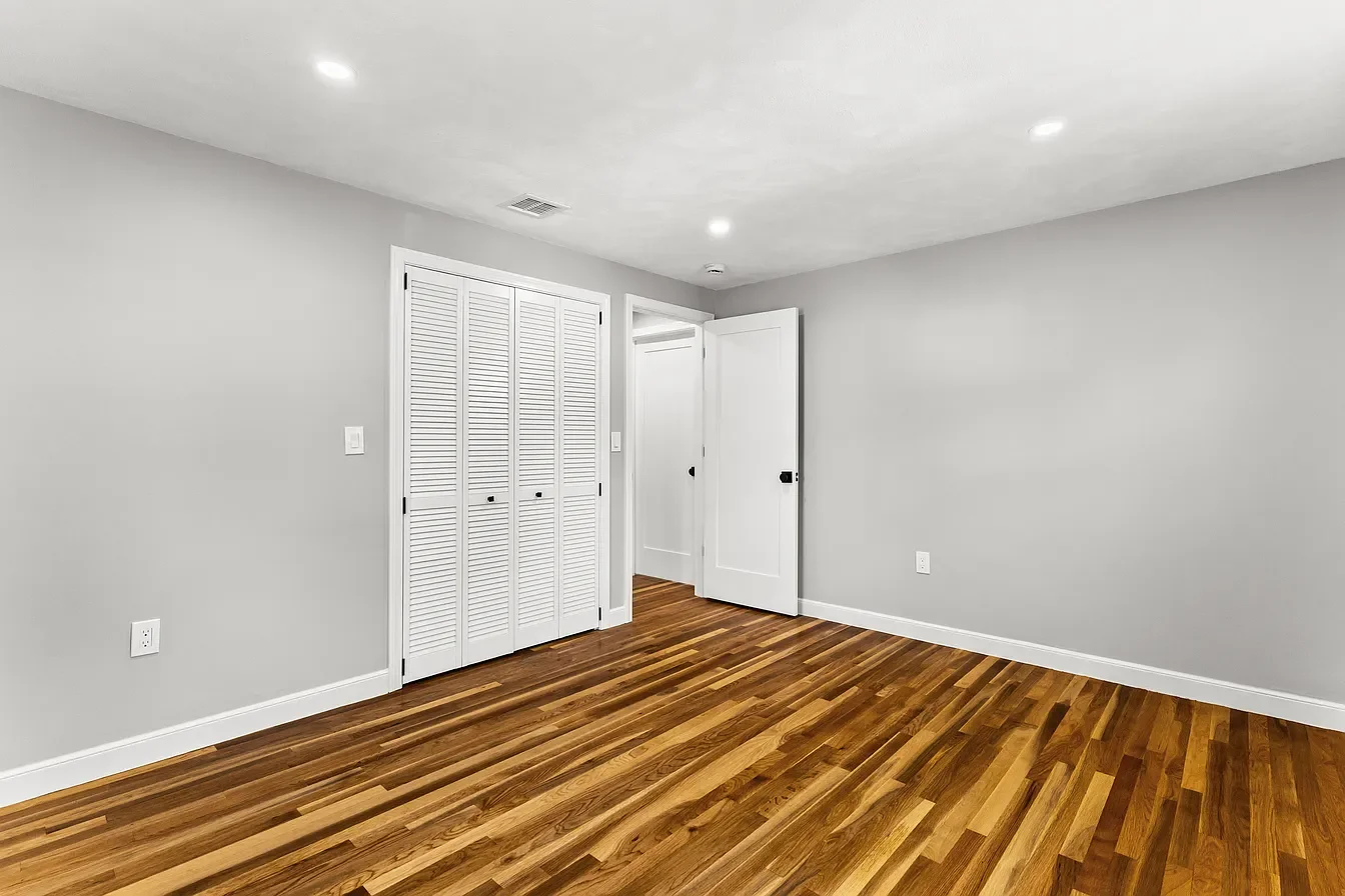 An empty room with light gray walls, hardwood flooring, a closet with white louvered doors, and an open door leading to another room. The ceiling has recessed lighting.