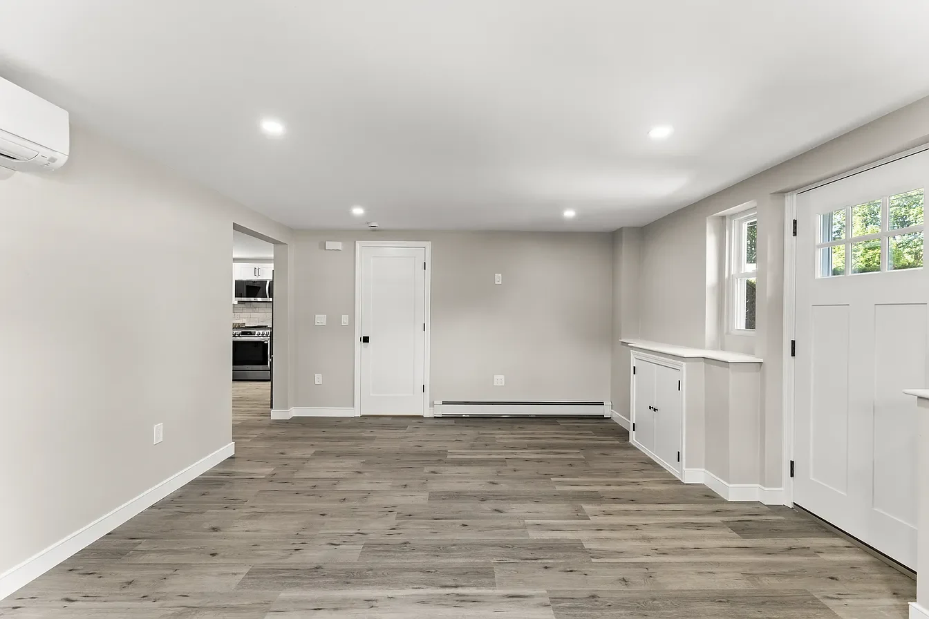 Empty living room with gray walls, wooden flooring, white trim, windows with blinds, and a white door leading to another room.