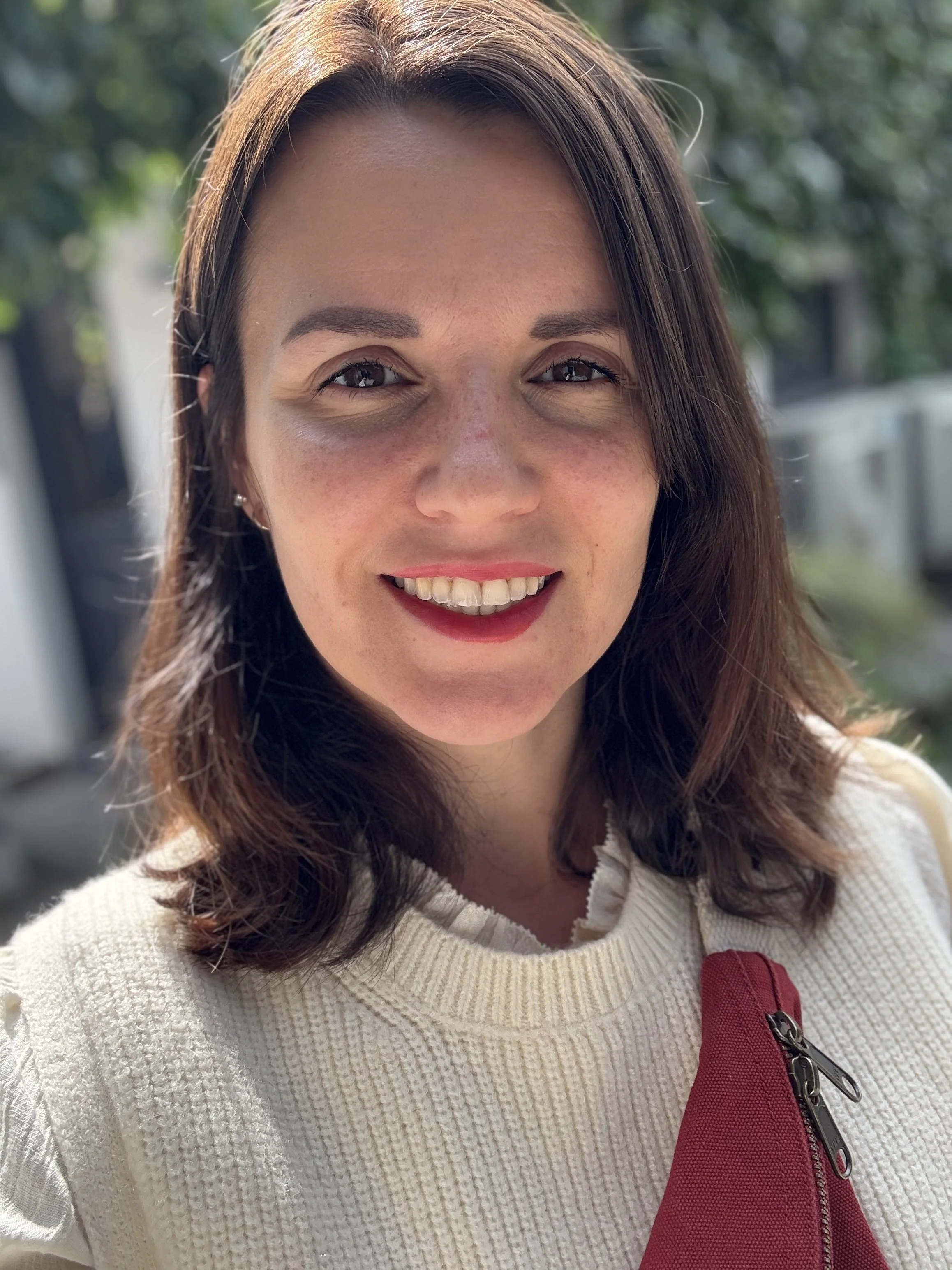 Jeune femme souriante, portant un pull blanc avec une fermeture éclair rouge, en extérieur avec des arbres en arrière-plan.