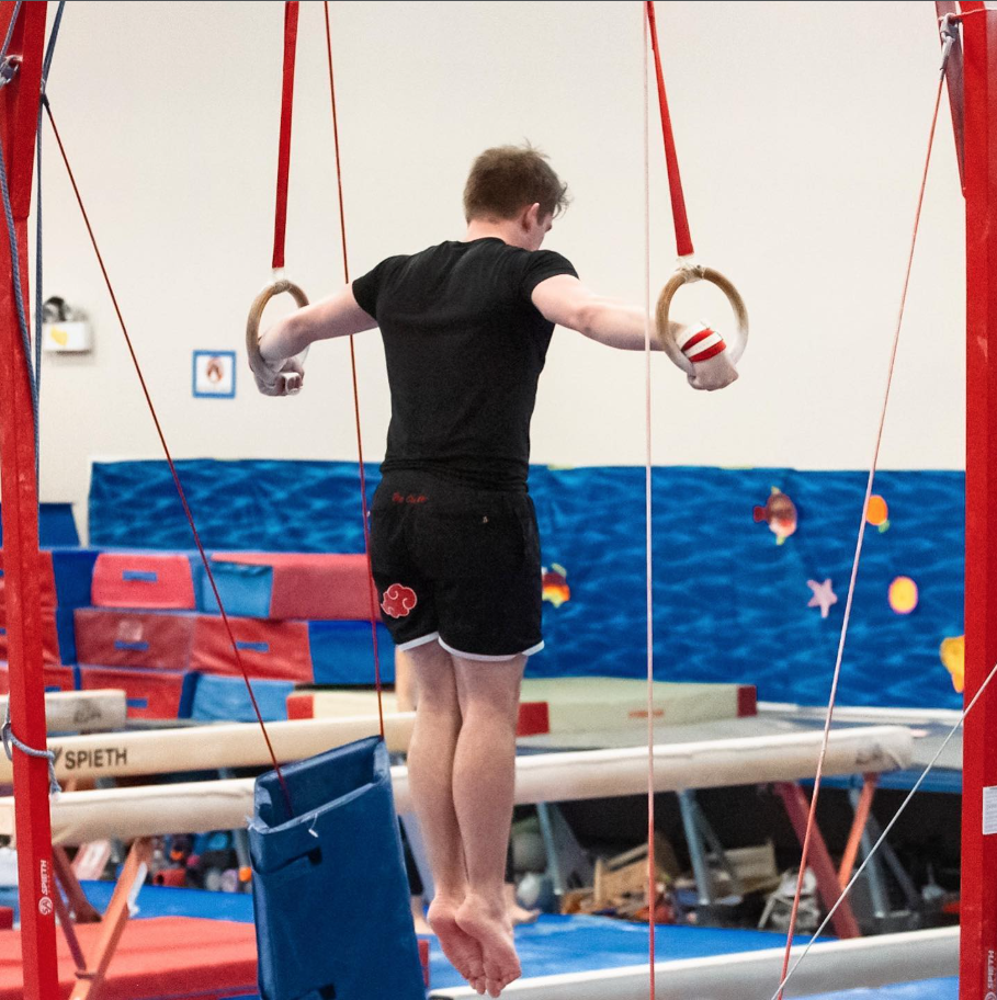 A person practicing on gymnastics rings in an indoor gym with colorful padded walls and gym equipment in the background.