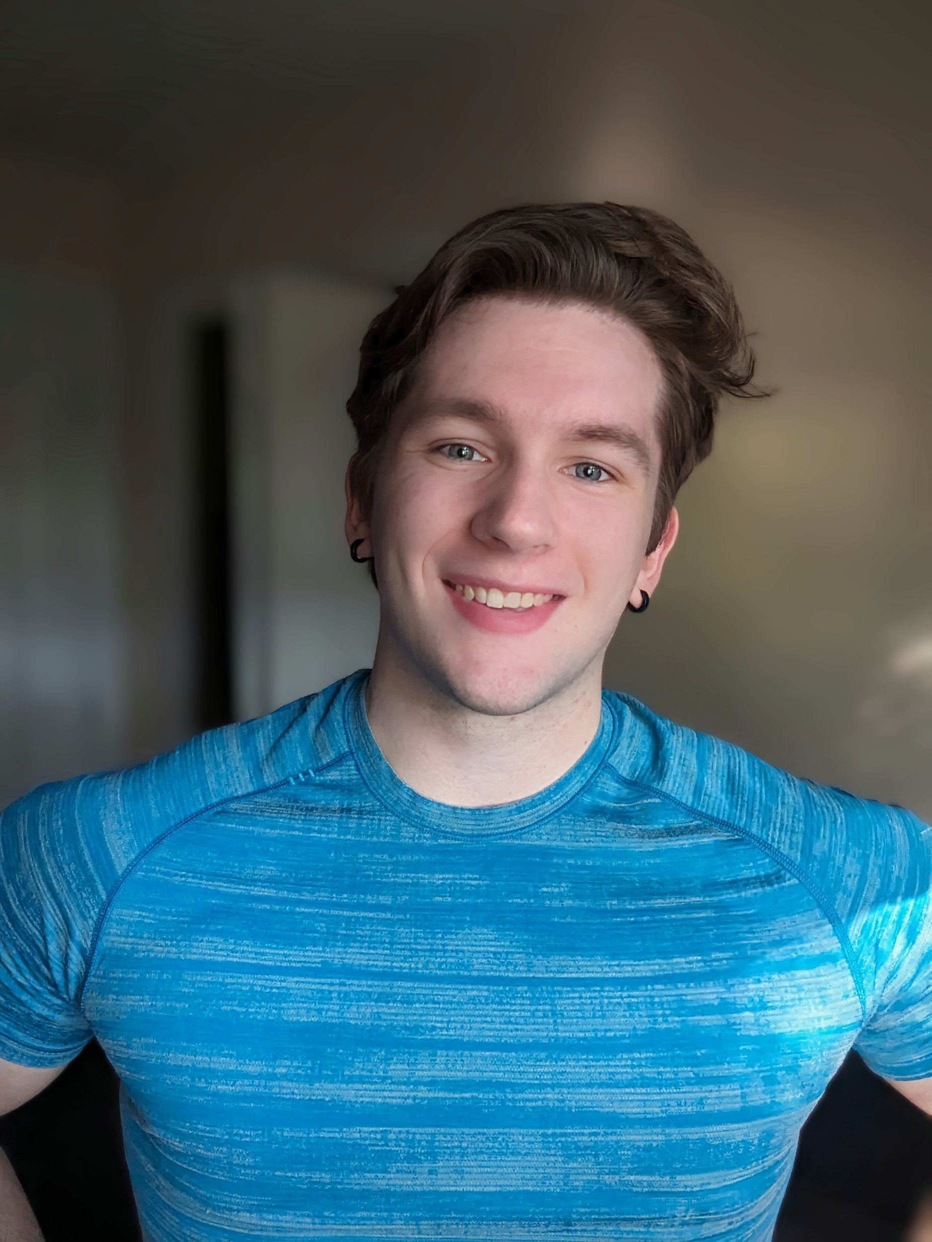 A young man with brown hair, blue eyes, and earrings, wearing a blue athletic shirt, smiling, in a room with a neutral background.