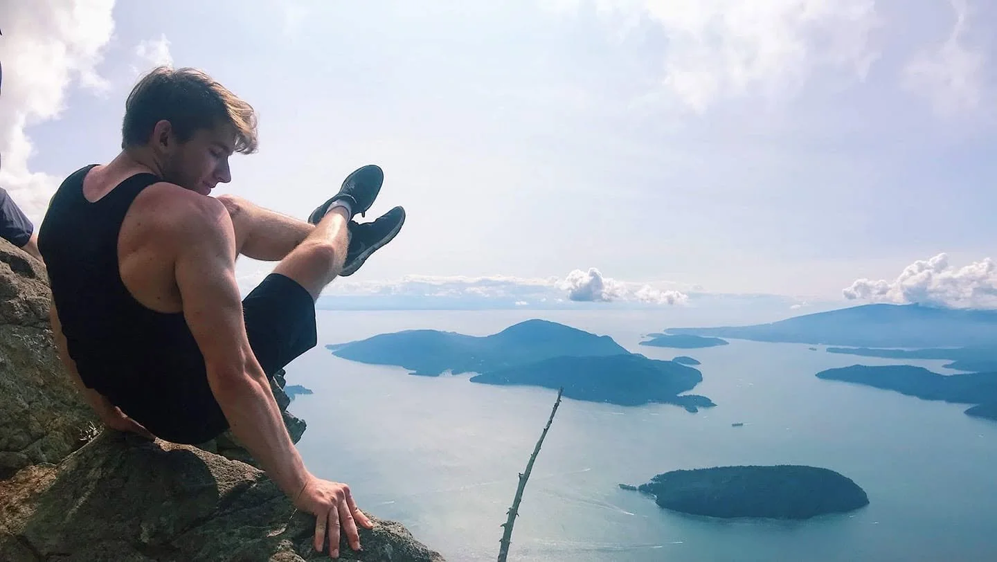 Person balancing on a rock ledge high above a body of water with islands in the distance.