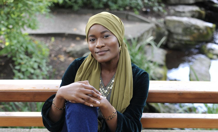 A woman with a beige head wrap and pearl earrings smiling outdoors near a stone archway with purple flowers