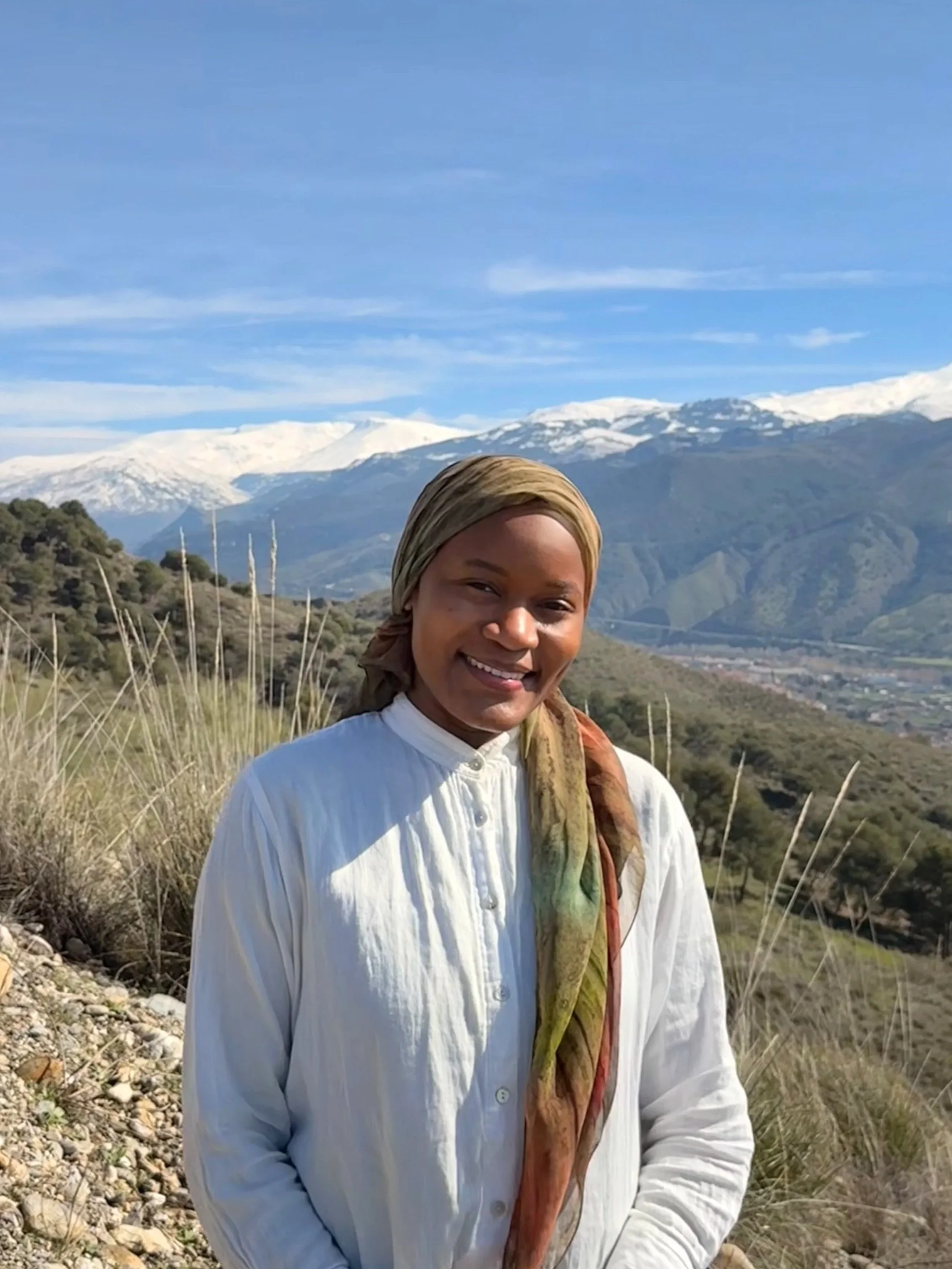 A woman with a multicolored head scarf and a white button down shirt smiling outdoors with a background of blue skies and snow-capped mountains.