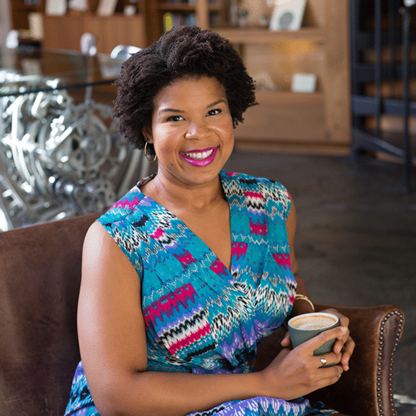A smiling woman with short curly hair and earrings, wearing a colorful patterned dress, sitting on a brown chair in a cozy cafe or coffee shop, holding a cup of coffee.