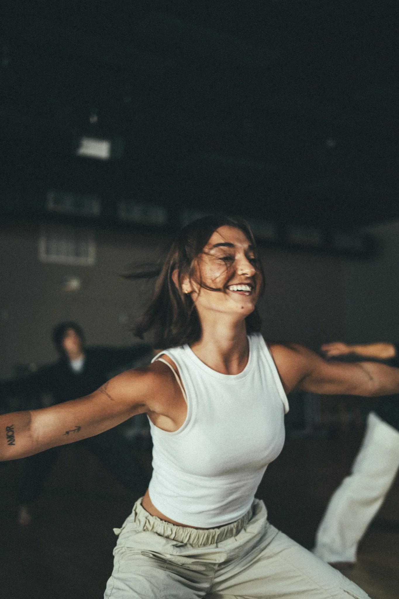 Woman with short brown hair dancing joyfully in a studio, wearing a white sleeveless top and beige pants, with other people in the background.