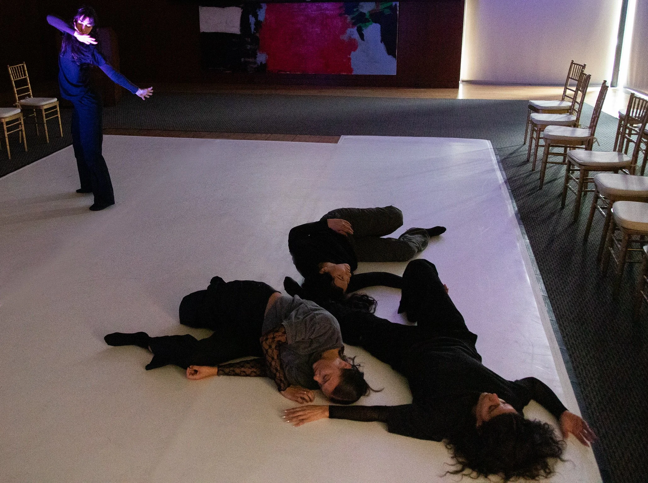 Four people are lying on the floor in a pattern resembling a star while a woman in a blue dress stands nearby, gesturing with her arm, inside a room with a white floor and chairs along the wall.
