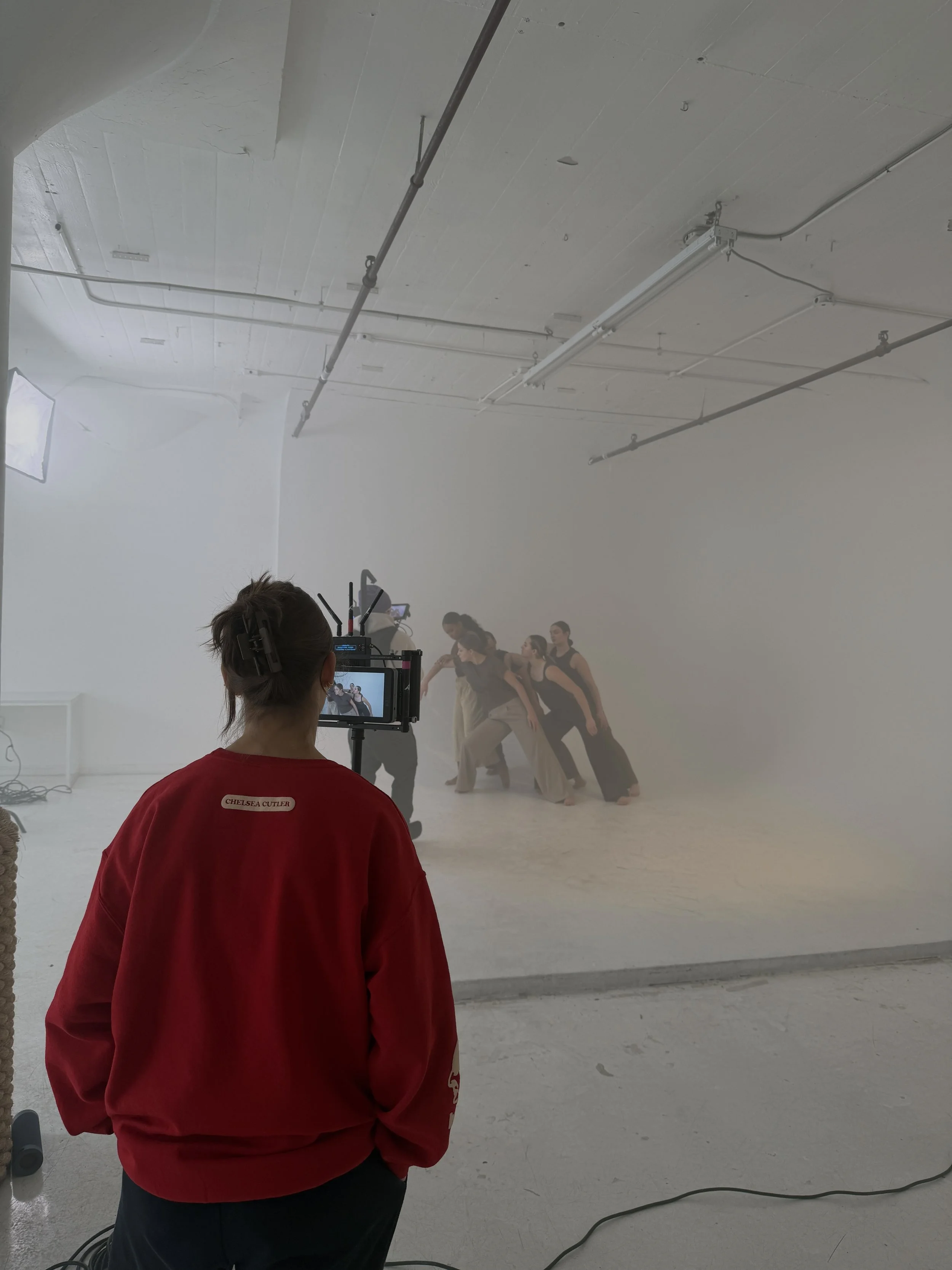 A behind-the-scenes view of a dance film recording in a white studio with five women dancing, wearing brown tops and pants, as gabby watches on the director's monitor.