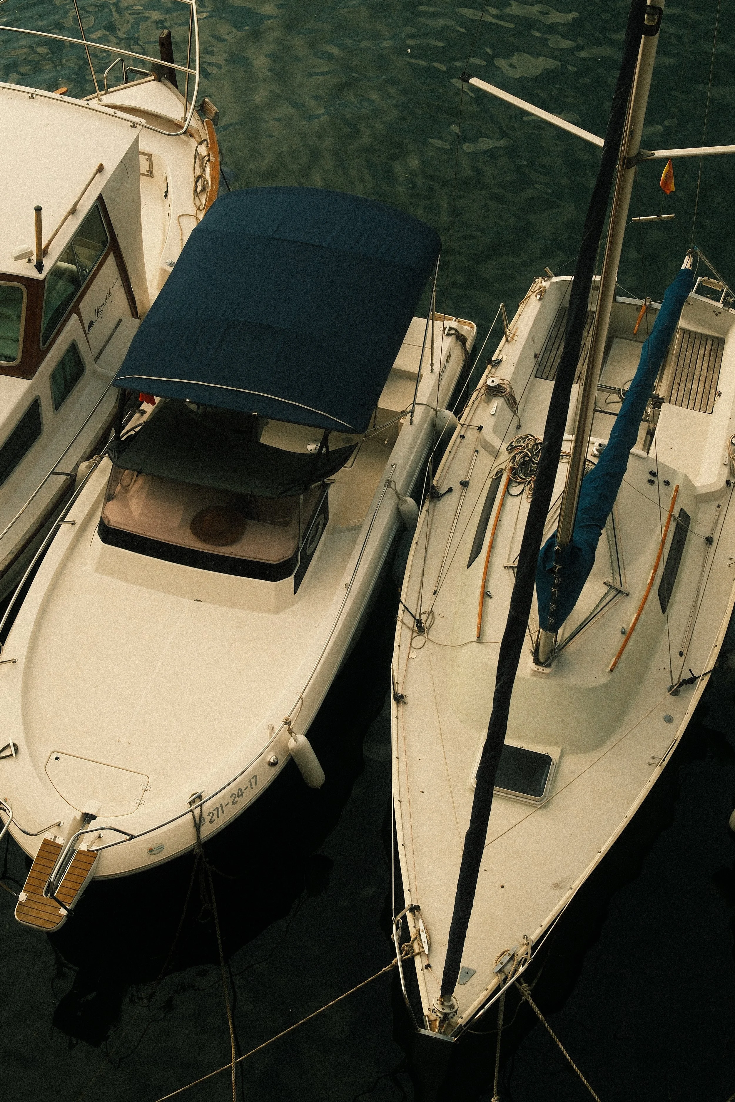 Top-down view of two docked boats on water, one sailboat on the right with a blue sail cover, and a motorboat on the left with a navy blue canopy.