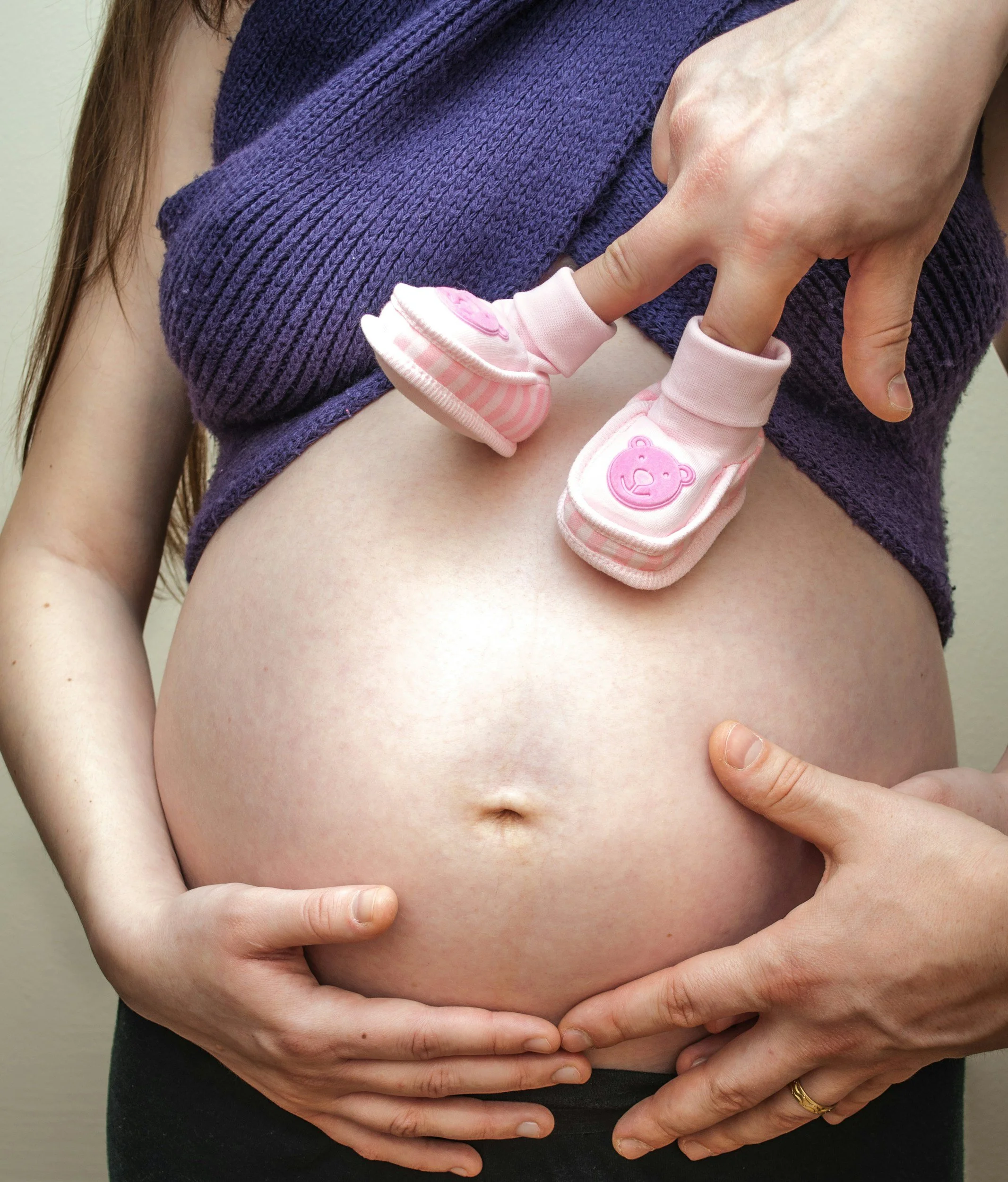 A pregnant person wearing a purple top, holding their bare belly with both hands. One hand is wearing a gold ring.