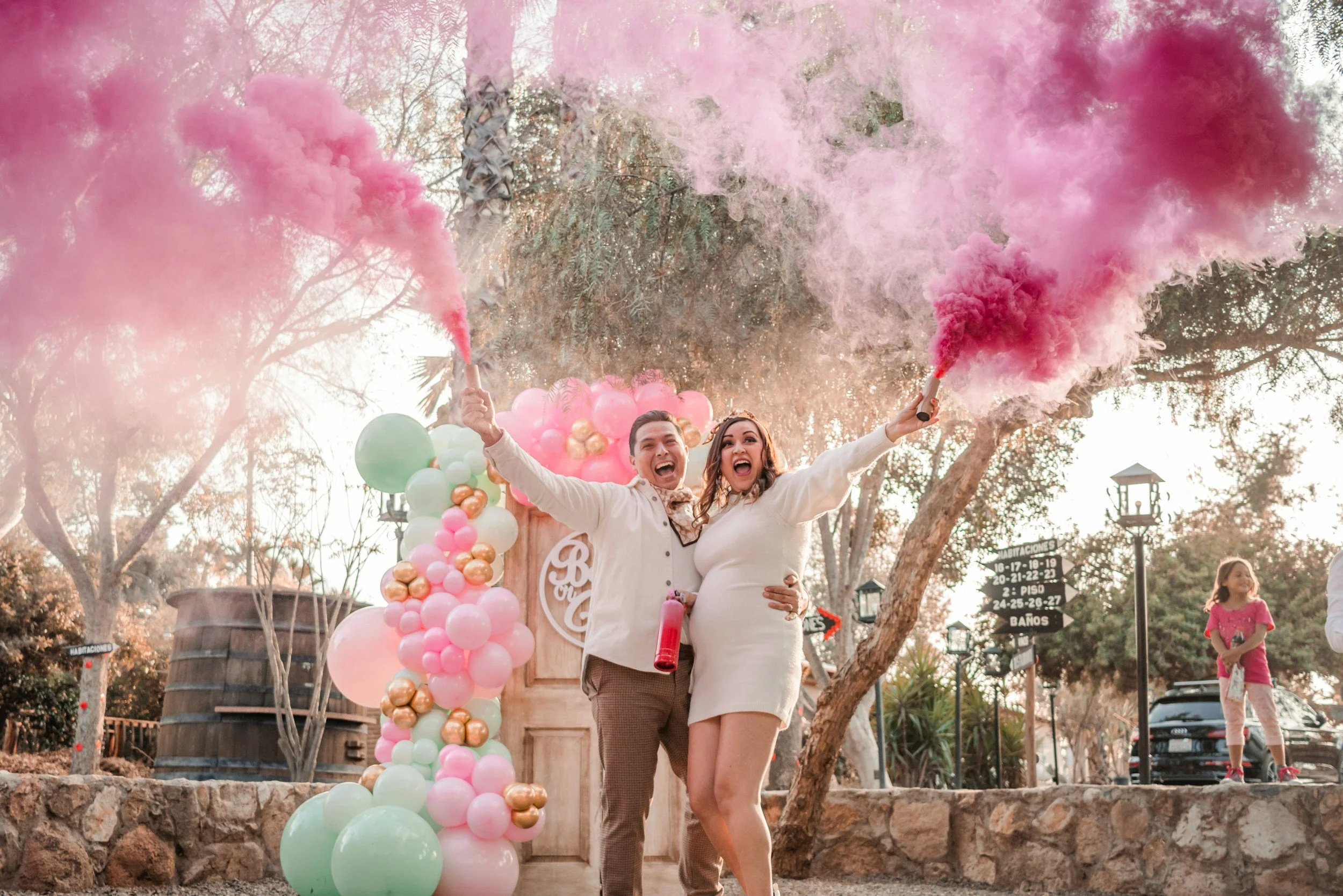 A couple celebrating with pink and gold balloons and smoke bombs outdoors, smiling with arms raised, surrounded by trees and festive decorations.