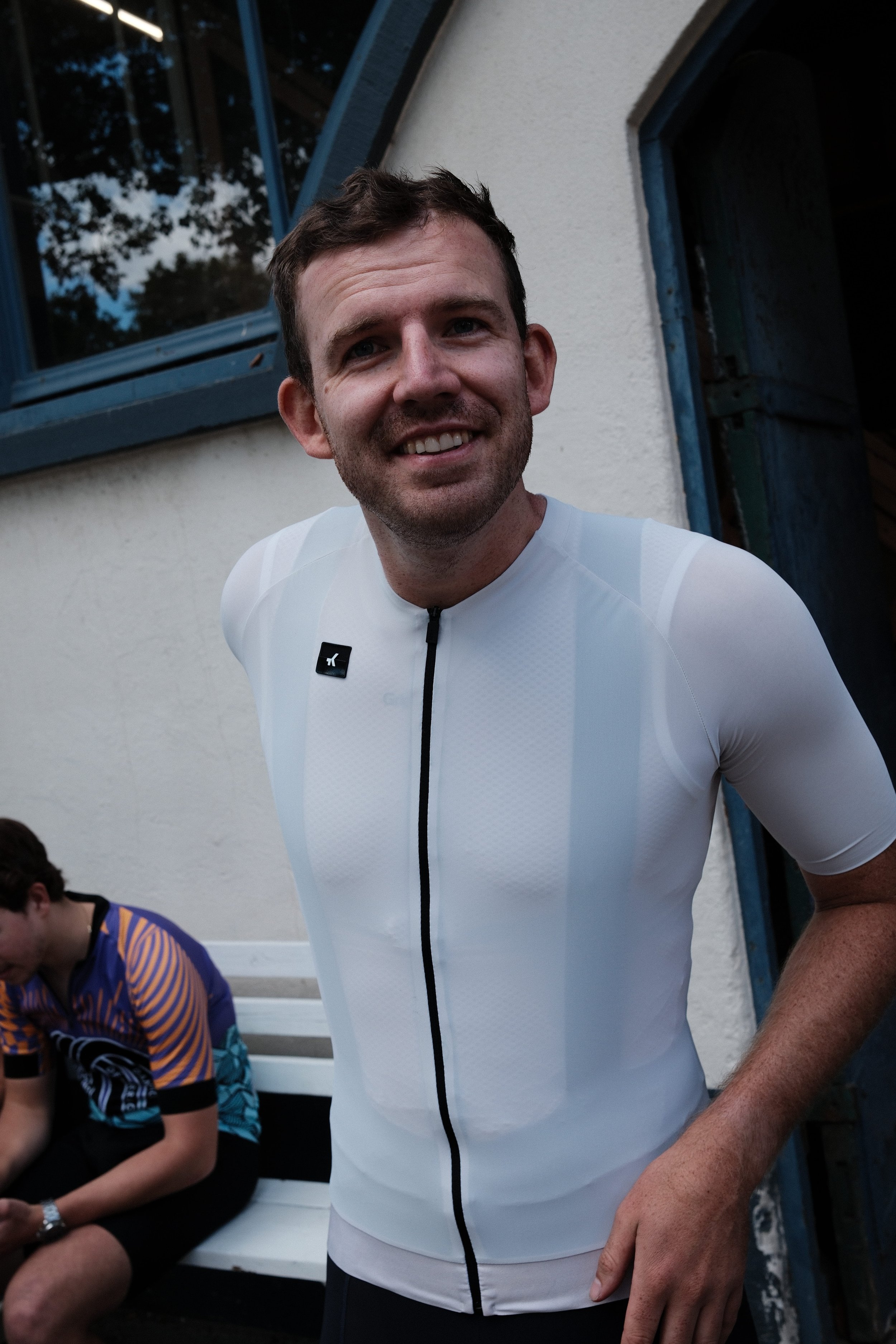 A smiling man in a white athletic cycling jersey standing outdoors next to a white building with a blue window frame, with a person in a colorful shirt sitting on a bench nearby.