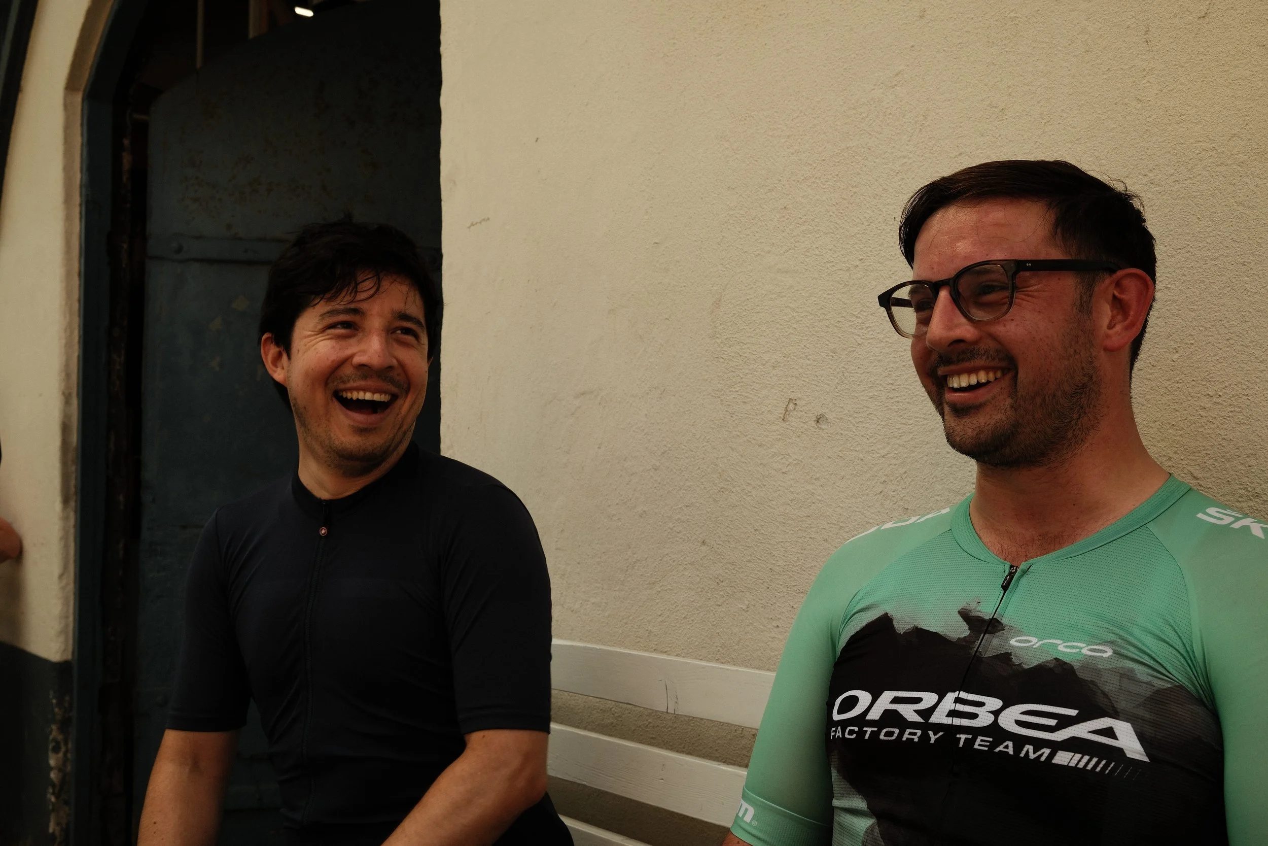 Two men are smiling and laughing, one wearing a black cycling jersey and the other wearing a green cycling jersey with glasses, sitting against a beige wall.