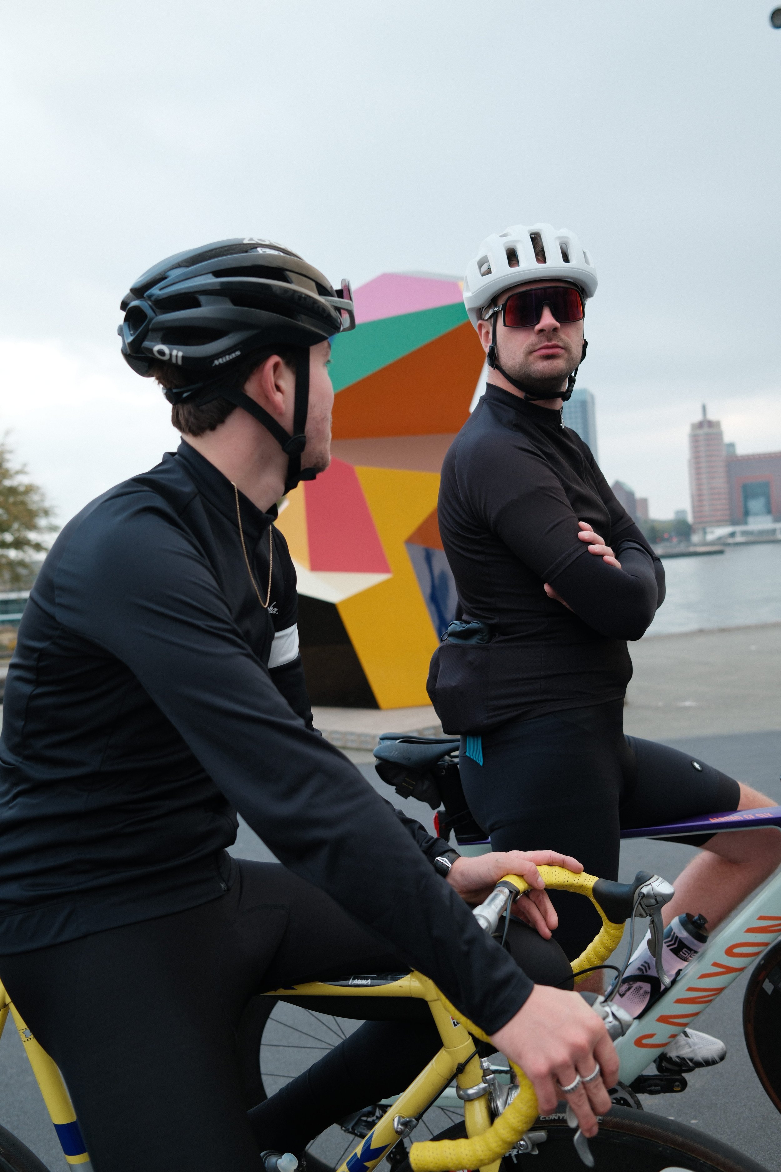 Two men in cycling gear and helmets, one sitting on a yellow Cannondale bike and the other standing with arms crossed, near colorful modern art sculpture by a river with city buildings in the background.