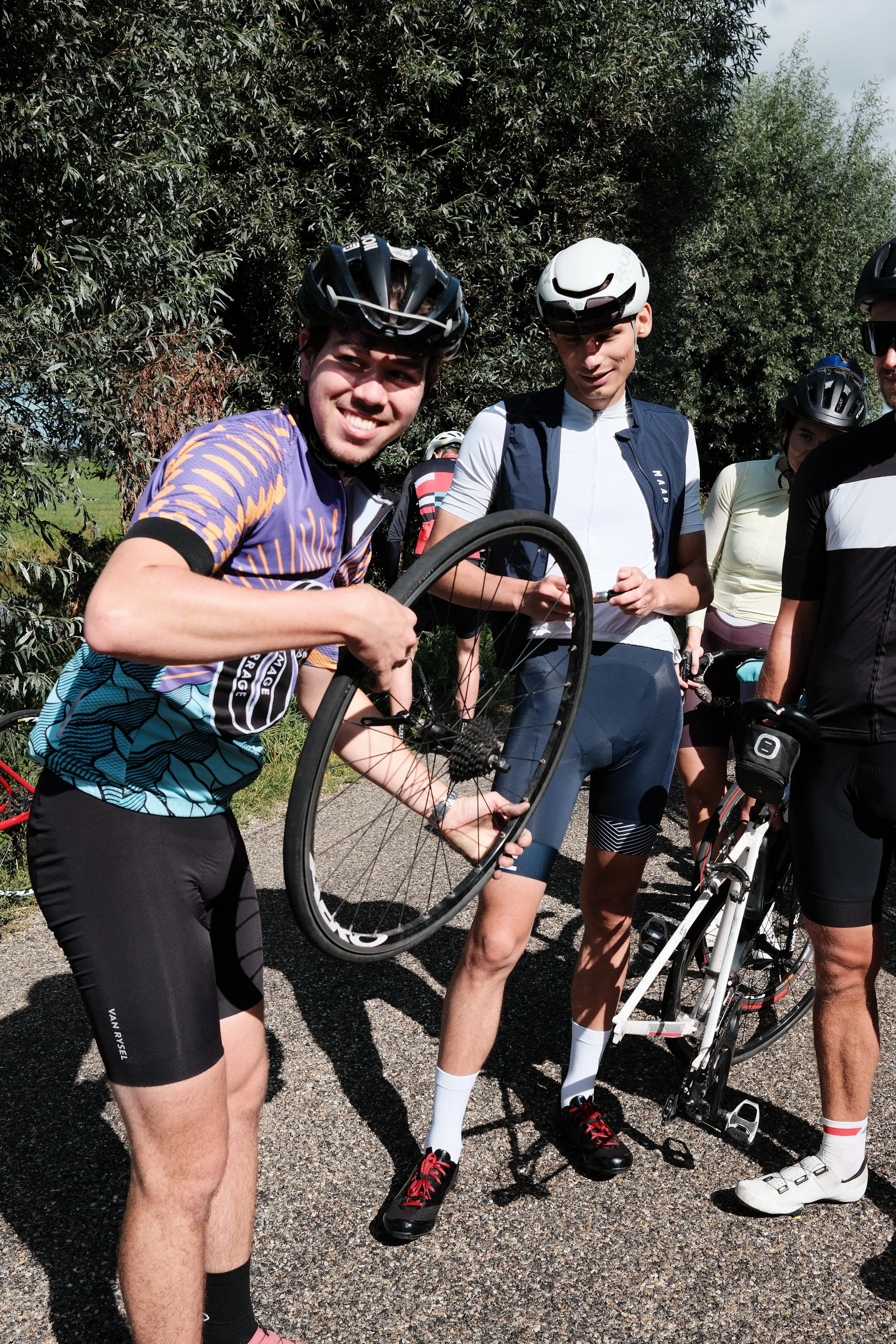 Group of cyclists in helmets and cycling gear standing outdoors on a gravel path with trees in the background.
