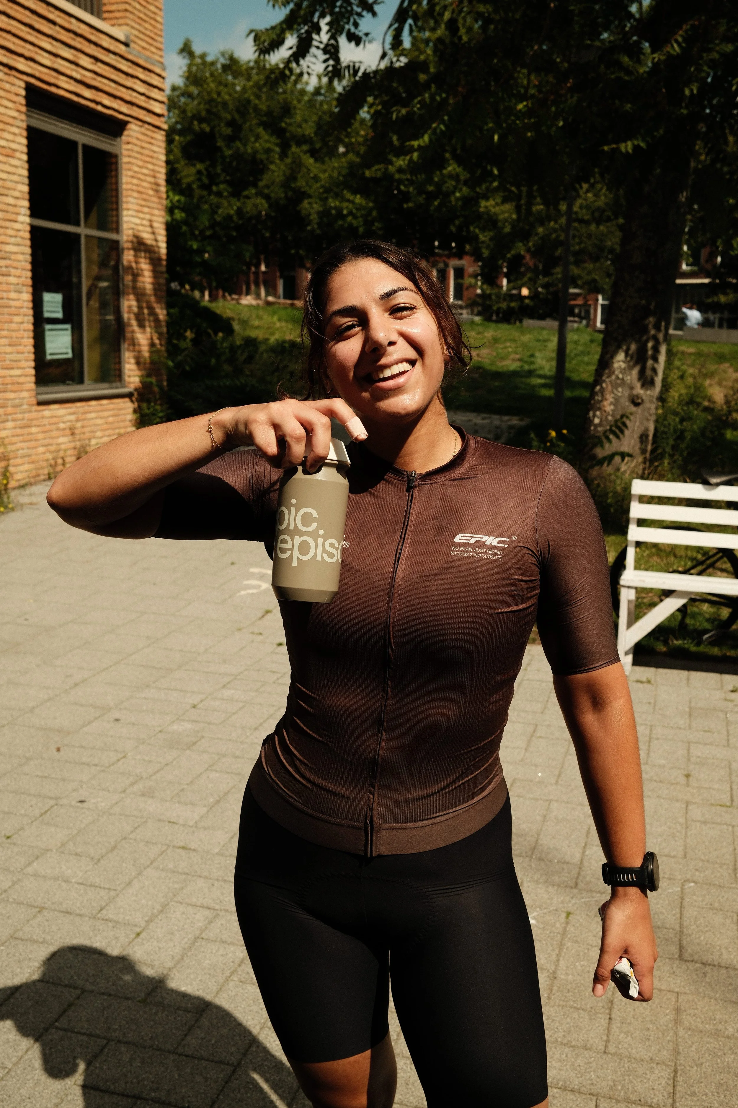 A young woman in athletic wear holding a water bottle and smiling outdoors on a sunny day.