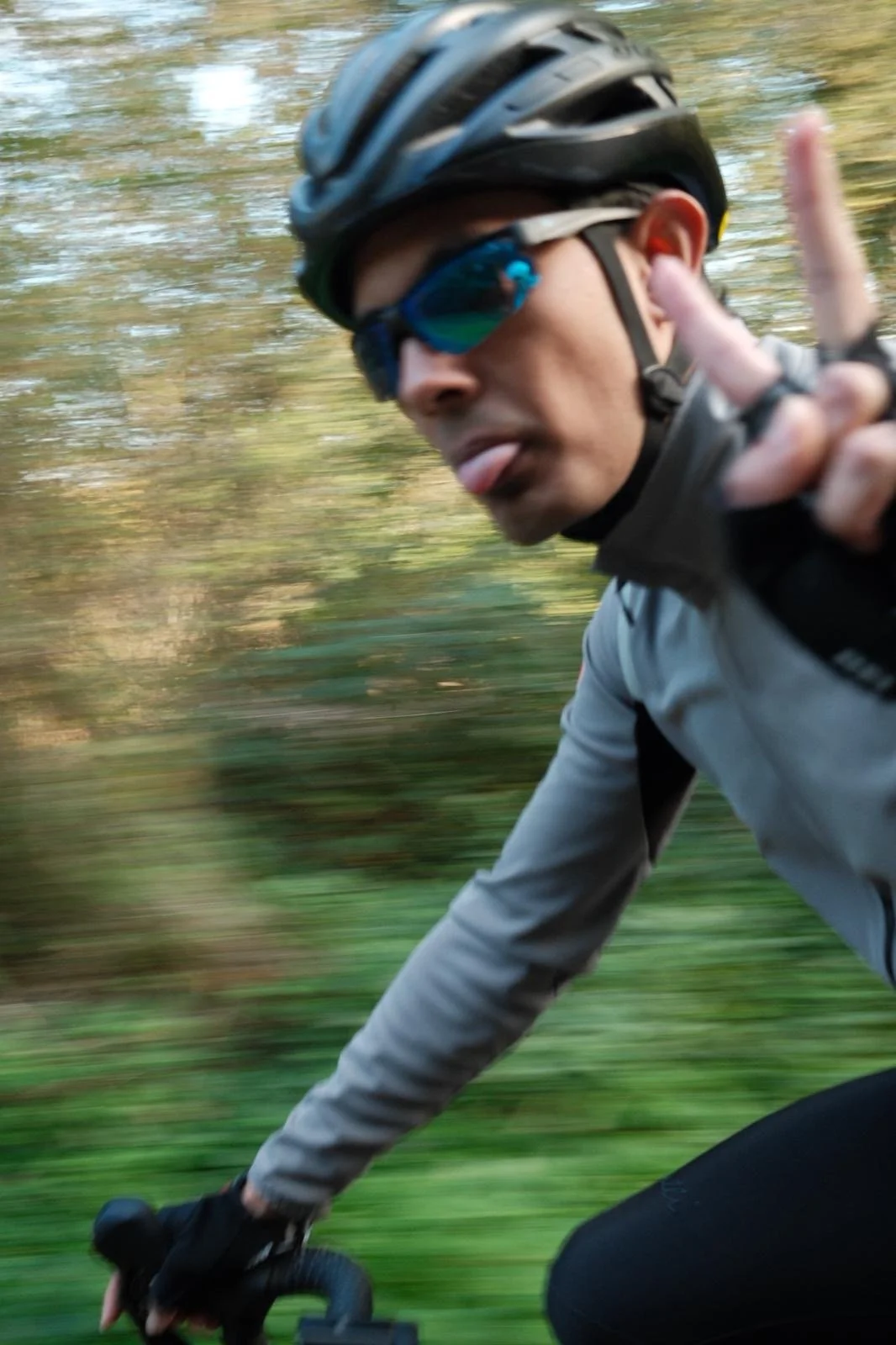 Cyclist wearing helmet and sunglasses sticking out tongue while riding a bike outdoors.