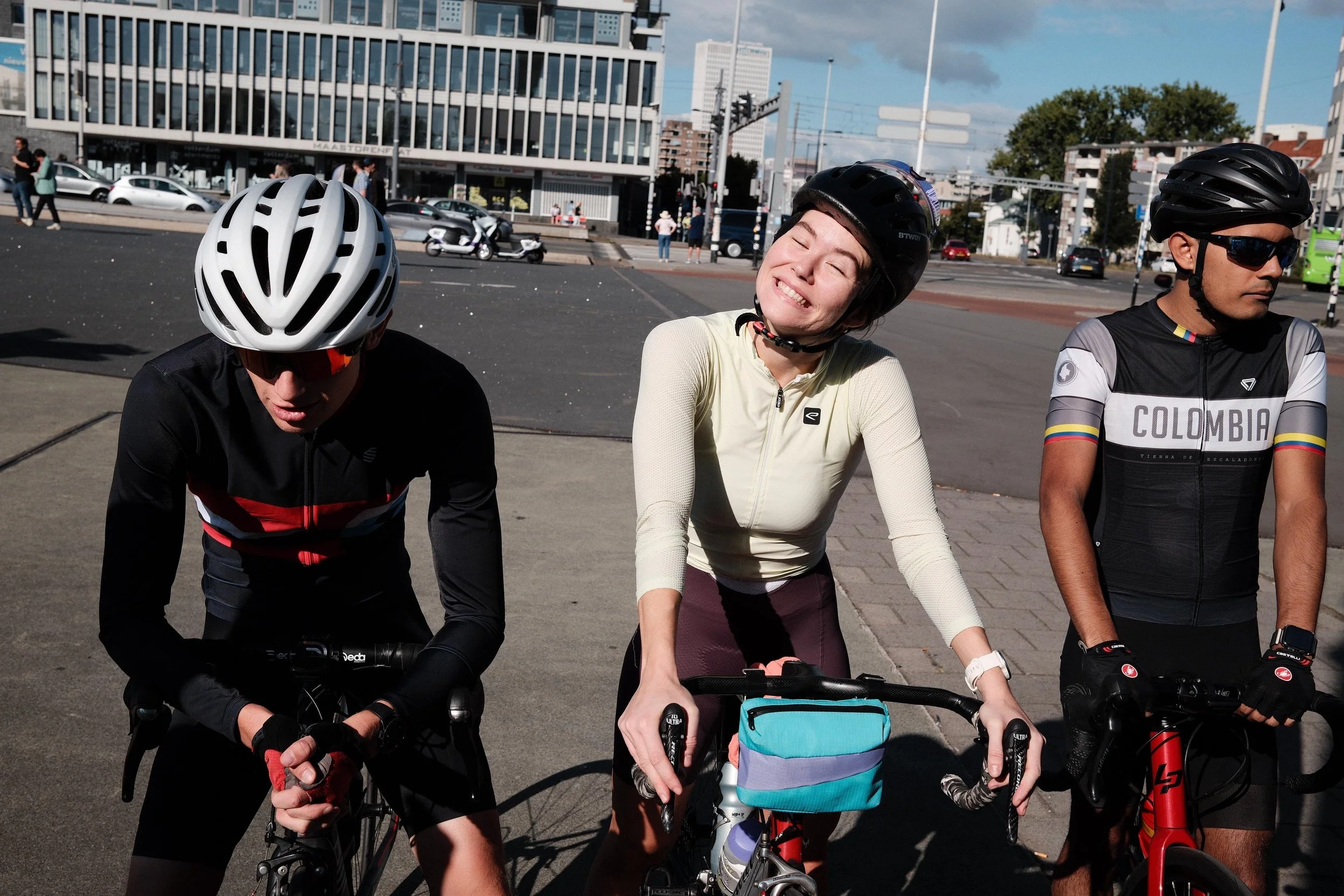 Three cyclists wearing helmets and cycling gear standing with their bikes on a city street, smiling and relaxing after a ride, with buildings and cars in the background.
