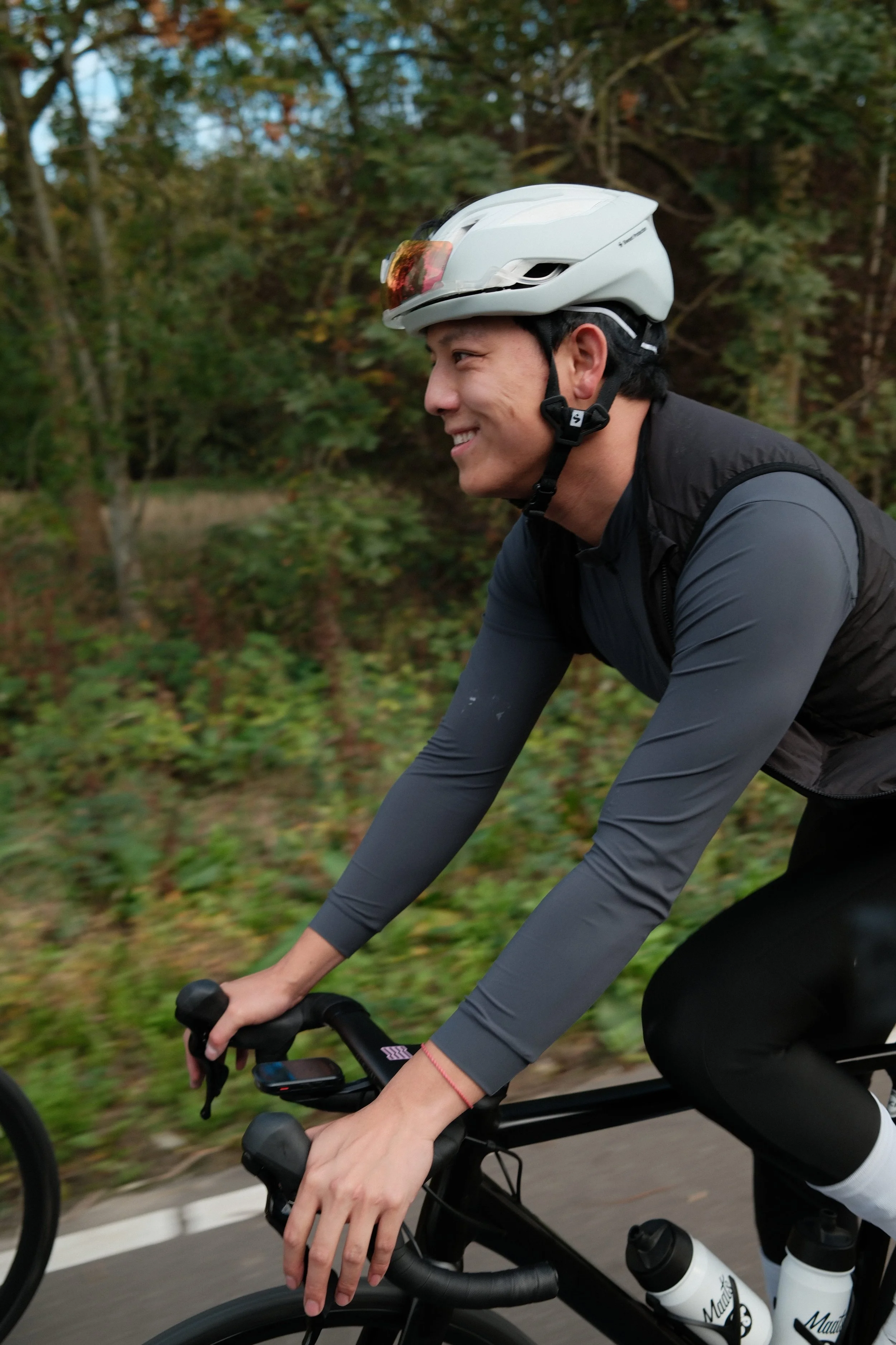 A man in cycling gear, including a helmet and sunglasses, riding a black road bike on a paved path surrounded by trees and greenery.