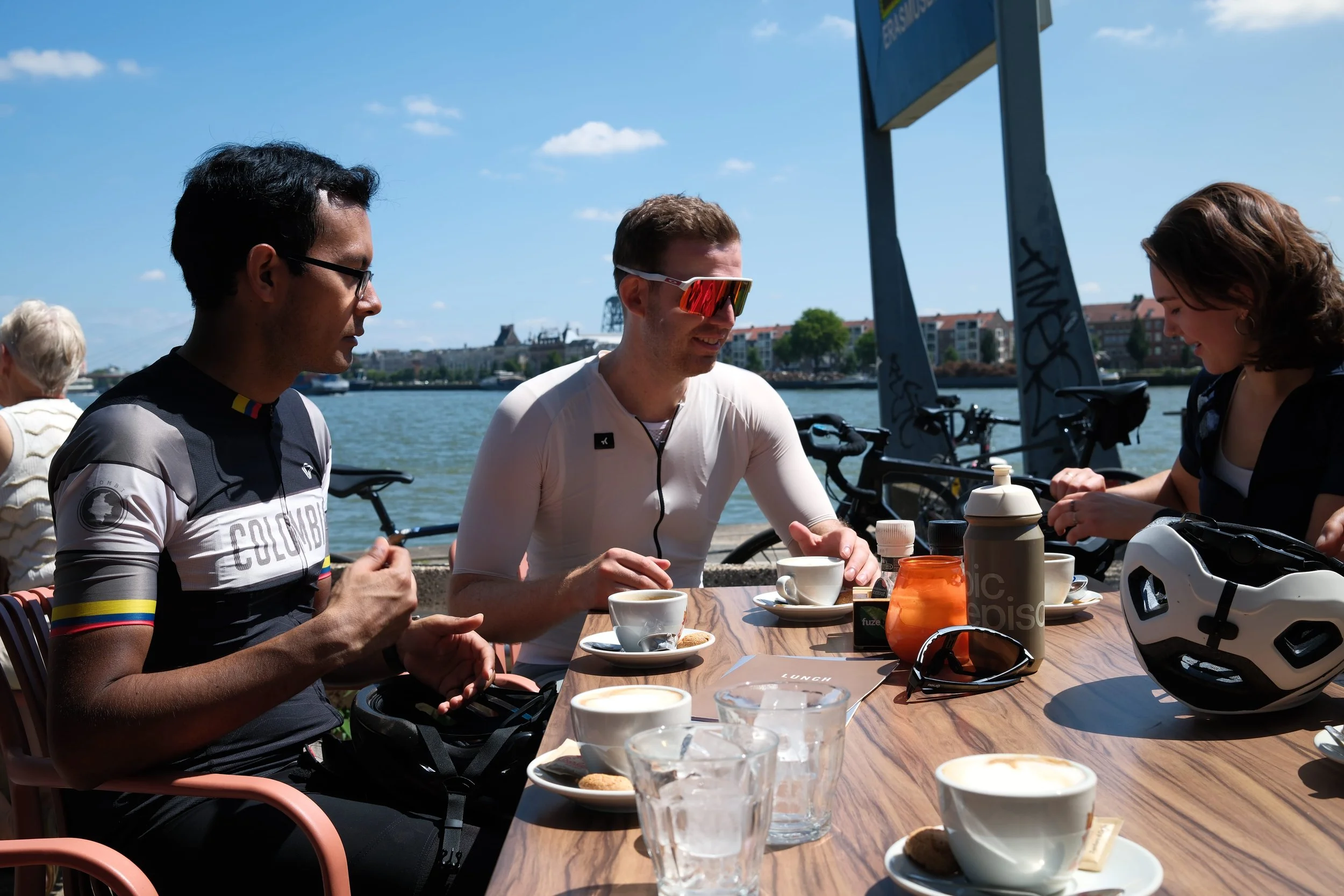 Three cyclists sitting at an outdoor table by a river, having coffee and snacks on a sunny day.