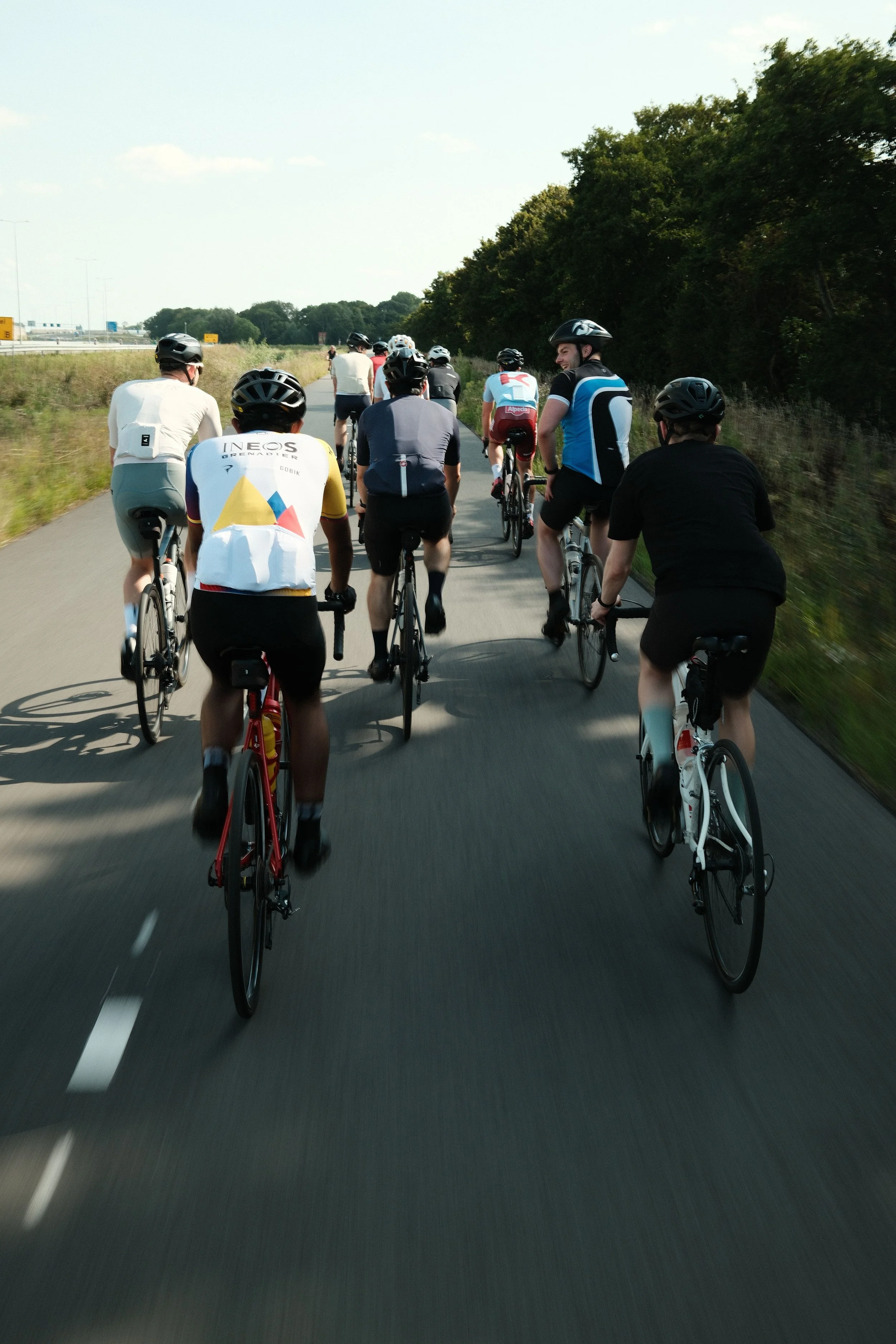 Group of people riding bicycles on a paved road surrounded by grassy fields and trees on a sunny day.
