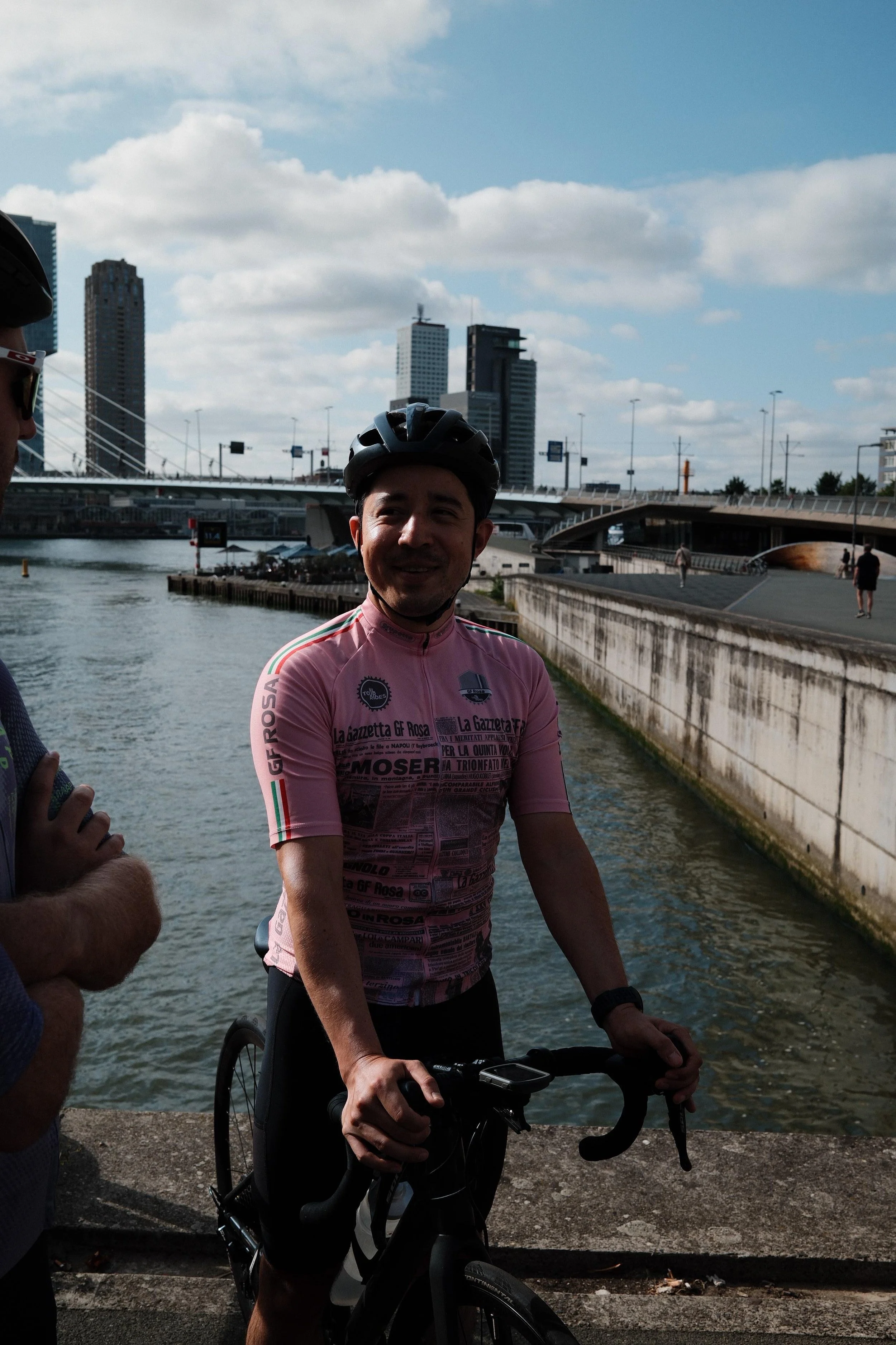 A man wearing a pink cycling jersey and a black helmet holding a bicycle by the water with a cityscape of tall buildings and a bridge in the background.