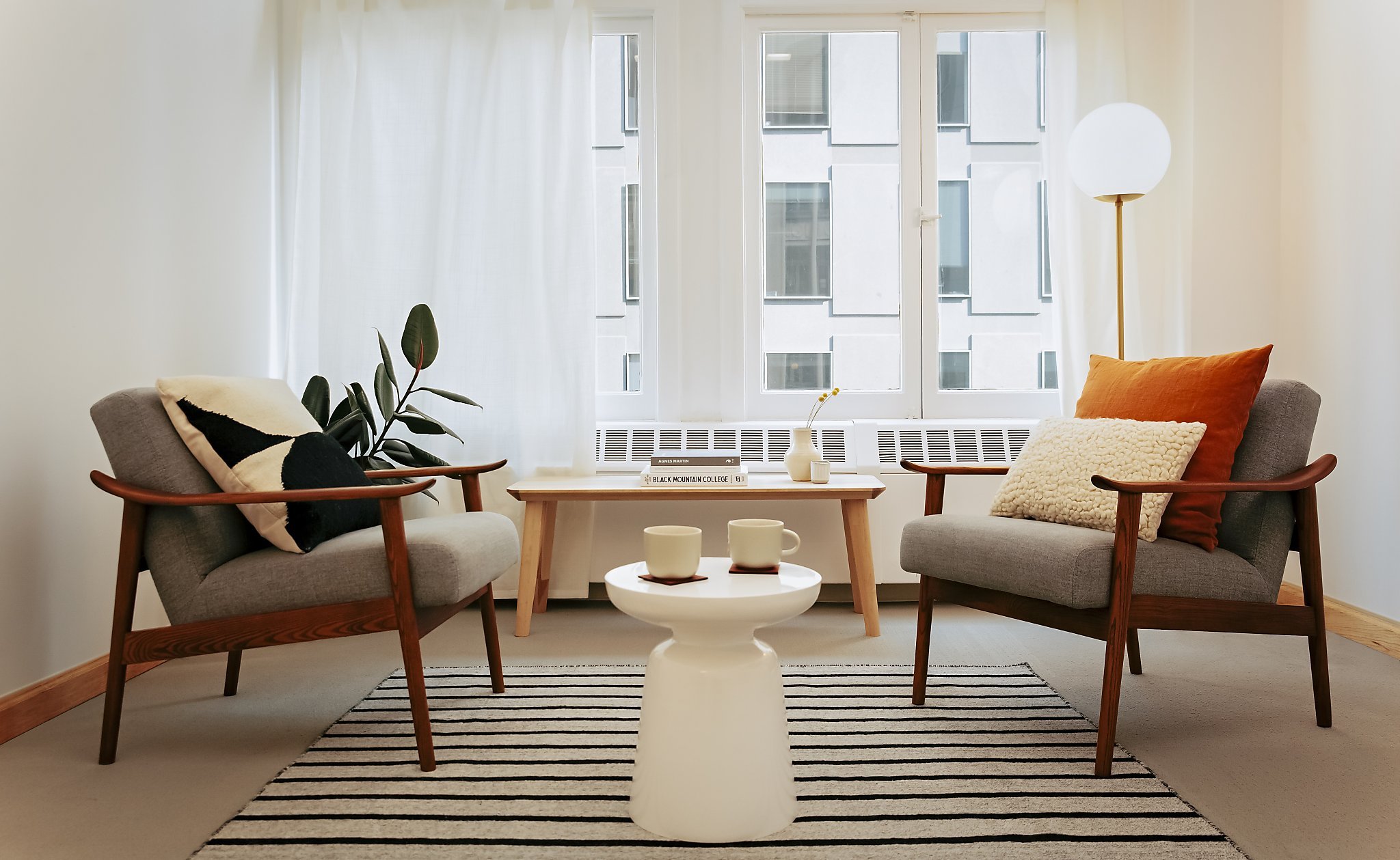 Living room with two gray armchairs with wooden arms, a white round side table with two ceramic mugs, a wooden coffee table with books and a white vase with a yellow flower, a striped rug, a large window with white curtains, a tall white lamp, and a potted plant.