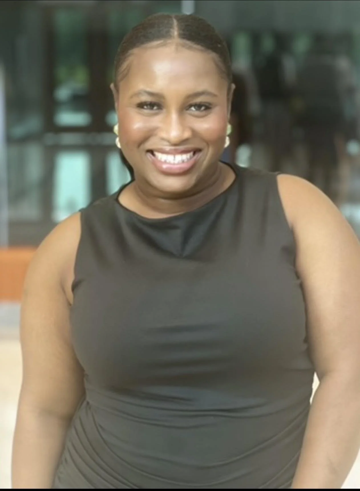 A smiling woman with dark hair styled back, wearing a sleeveless black top, standing indoors with a blurred background.
