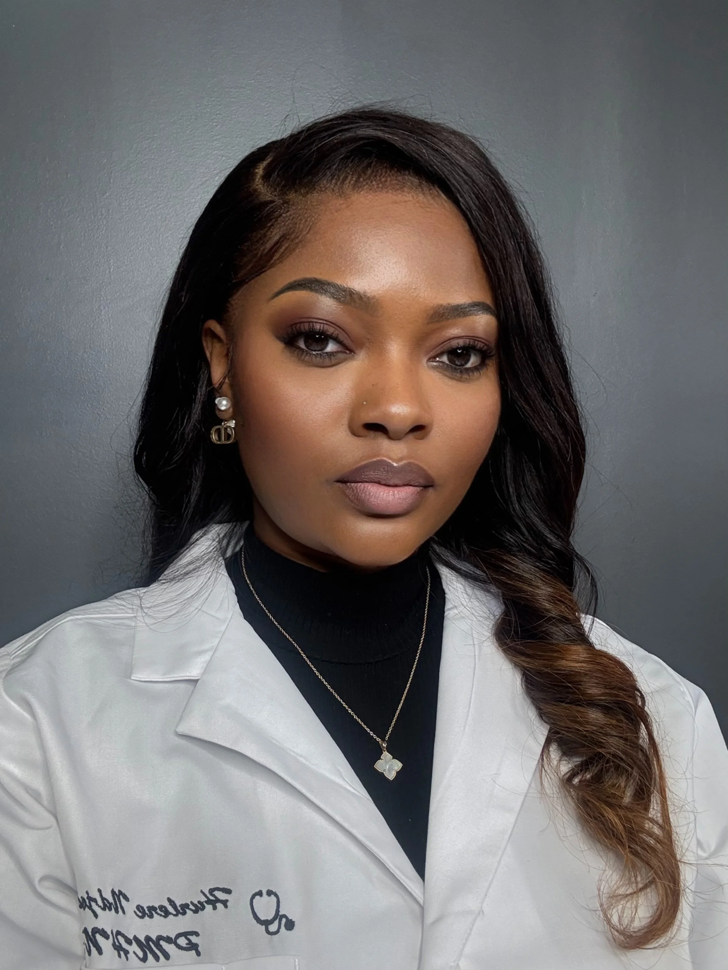 A professional woman with dark hair and brown skin, wearing makeup, earrings, a necklace, a black turtleneck, and a white coat with embroidered text, standing against a dark gray background.