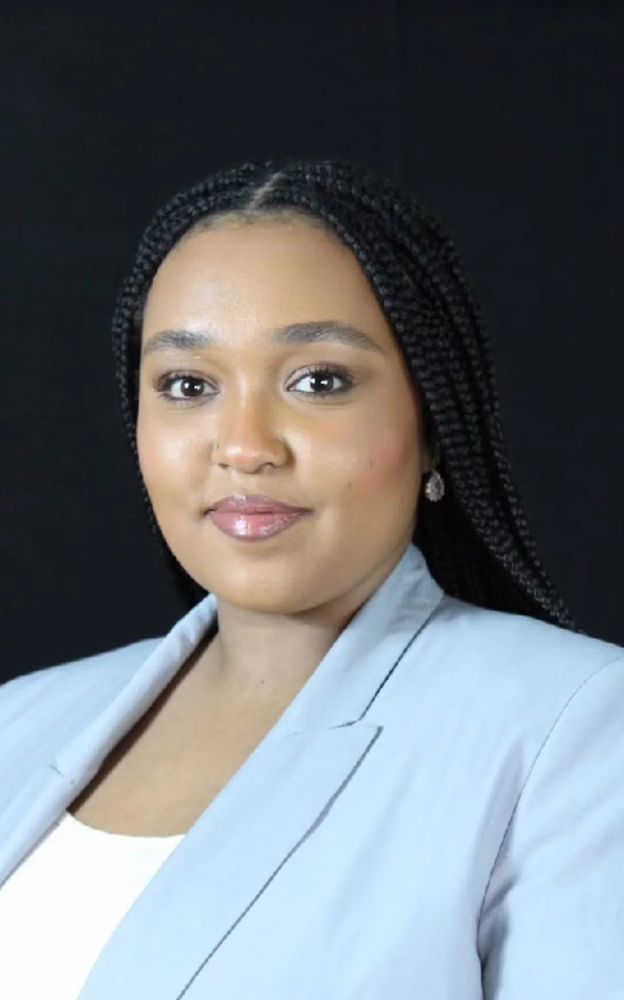A professional woman with braided hair, wearing a light gray blazer and a white top, posing against a black background.