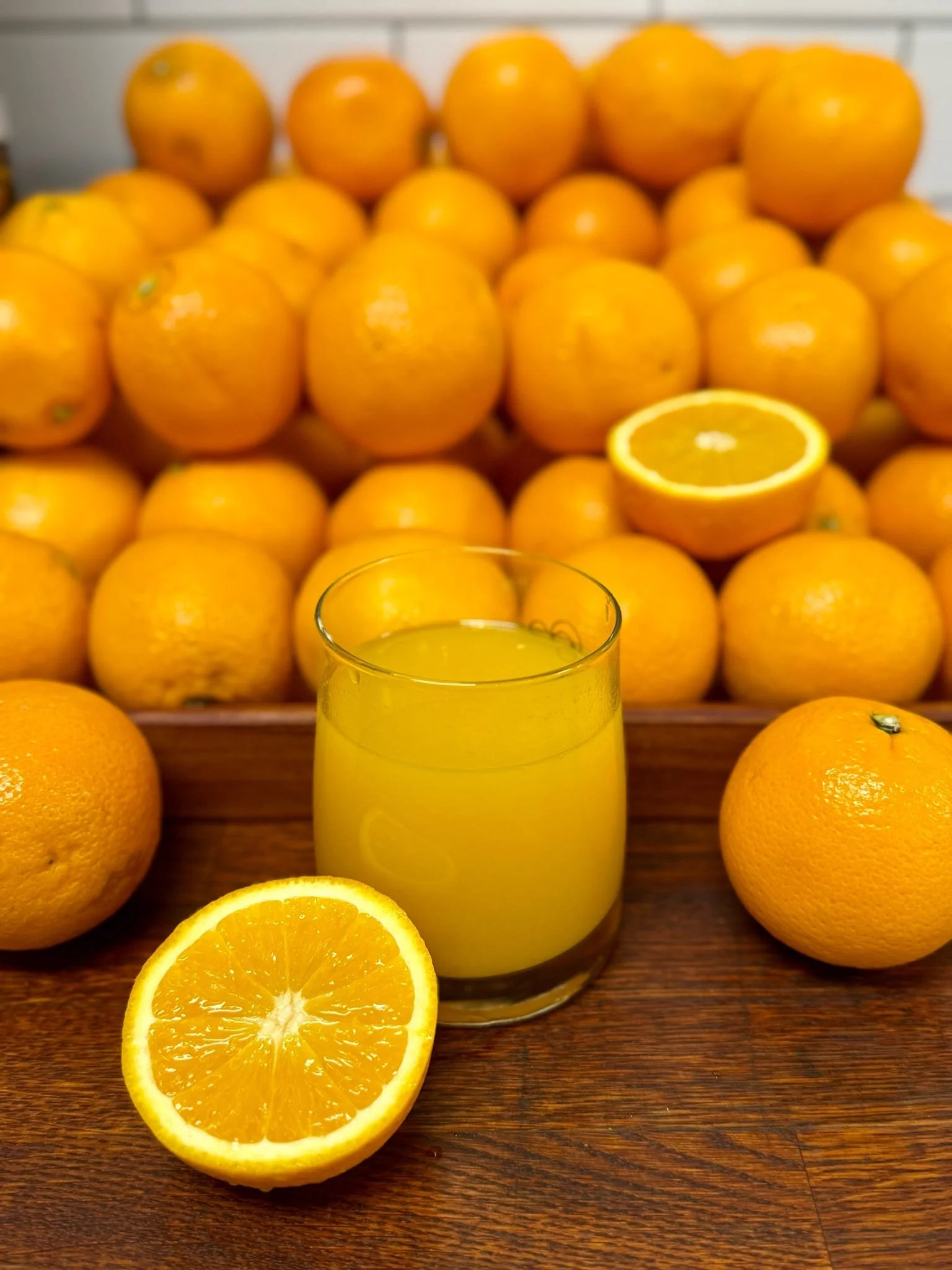 A glass of orange juice in front of a halved orange and a pile of oranges in the background.