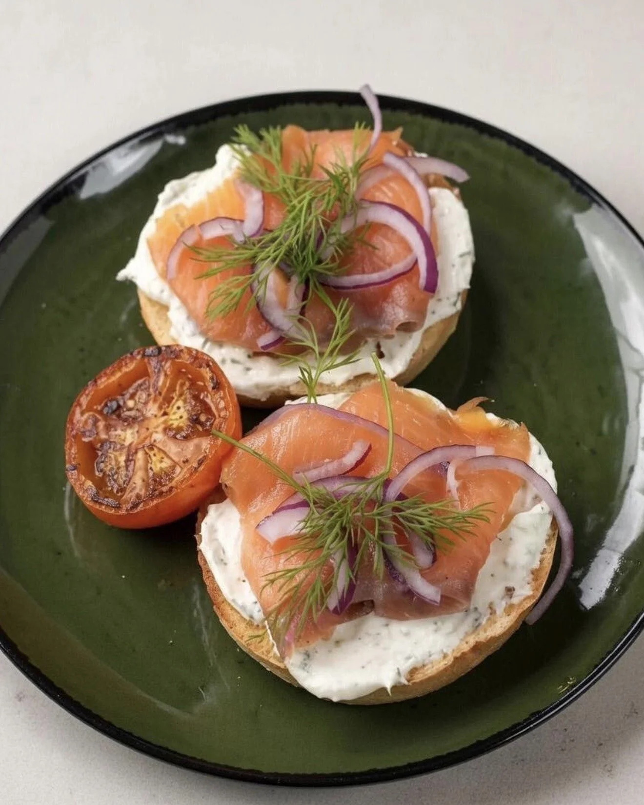 Two toasted bagel halves topped with cream cheese, smoked salmon, red onion slices, and garnished with fresh dill on a green plate.