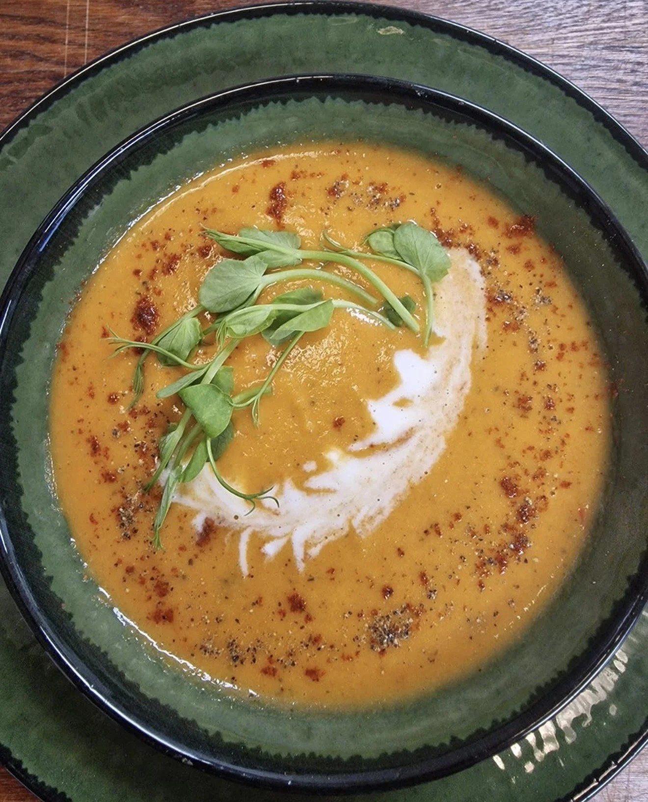 A bowl of creamy orange soup garnished with fresh green herbs and a swirl of cream, on a dark green plate.