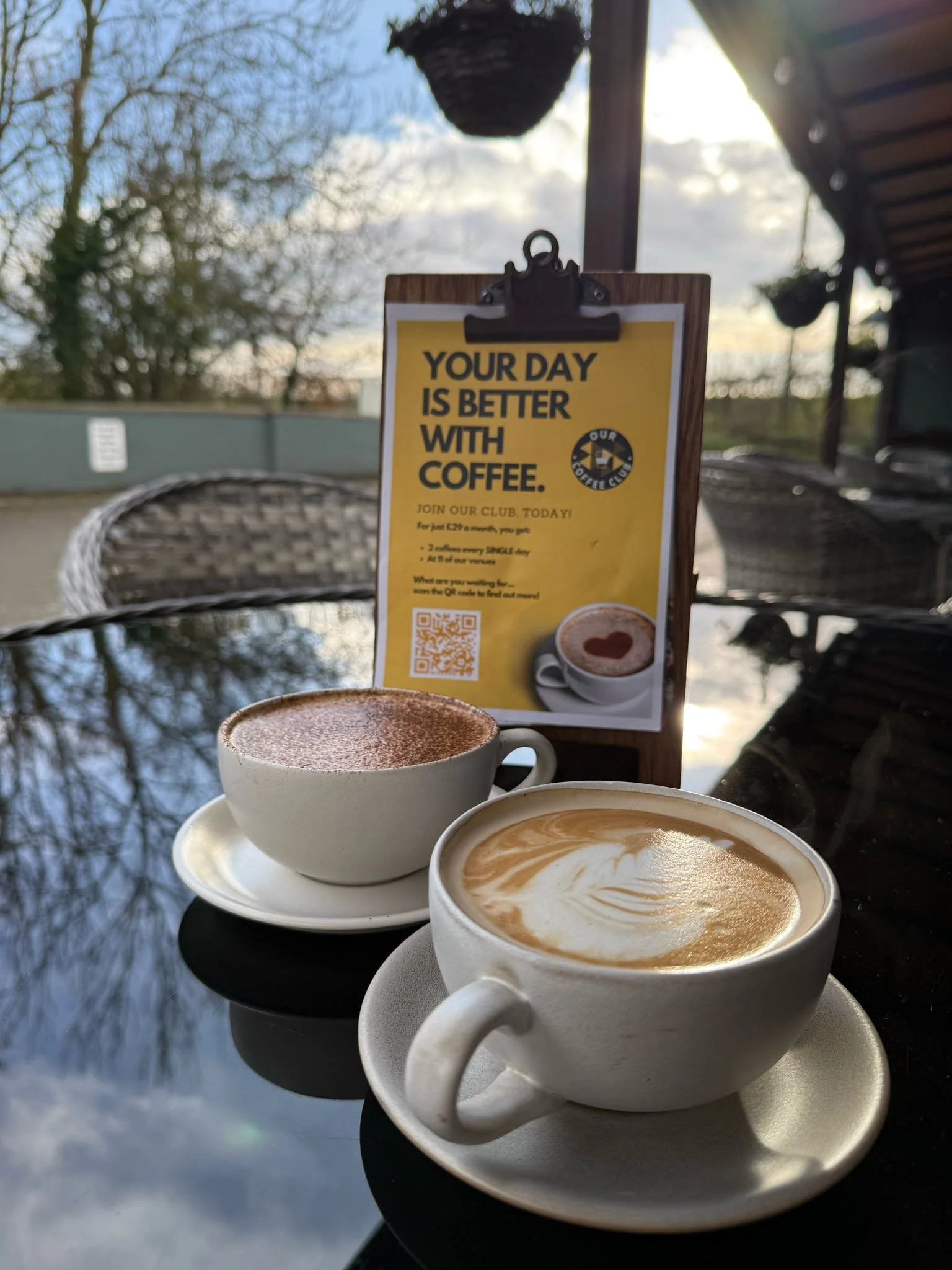 Two cups of coffee on a table outdoors, a promotional sign in the background with a yellow section offering coffee club membership, and a scenic outdoor setting with trees and a cloudy sky.